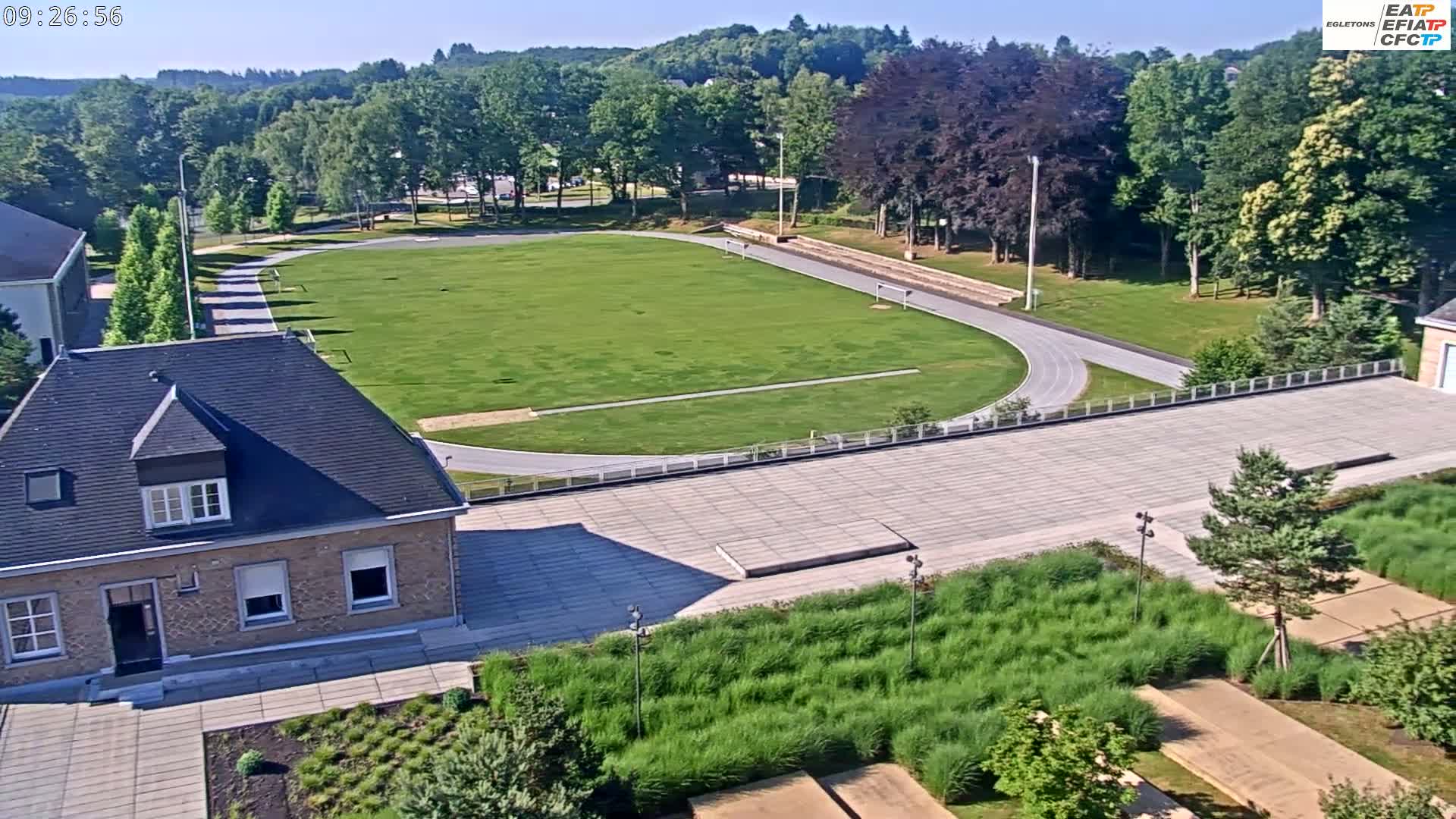 An aerial view shows a grassy athletic field with a running track, a small brick building, and surrounding landscaping on a sunny day.