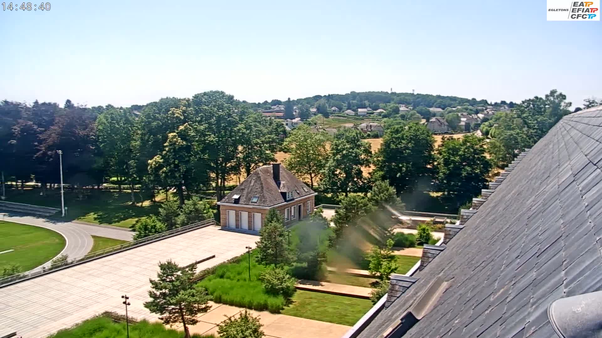 A sunny view overlooking a courtyard with a small brick building, a running track, and lush green landscaping, all bordered by trees and a small village in the distance.
