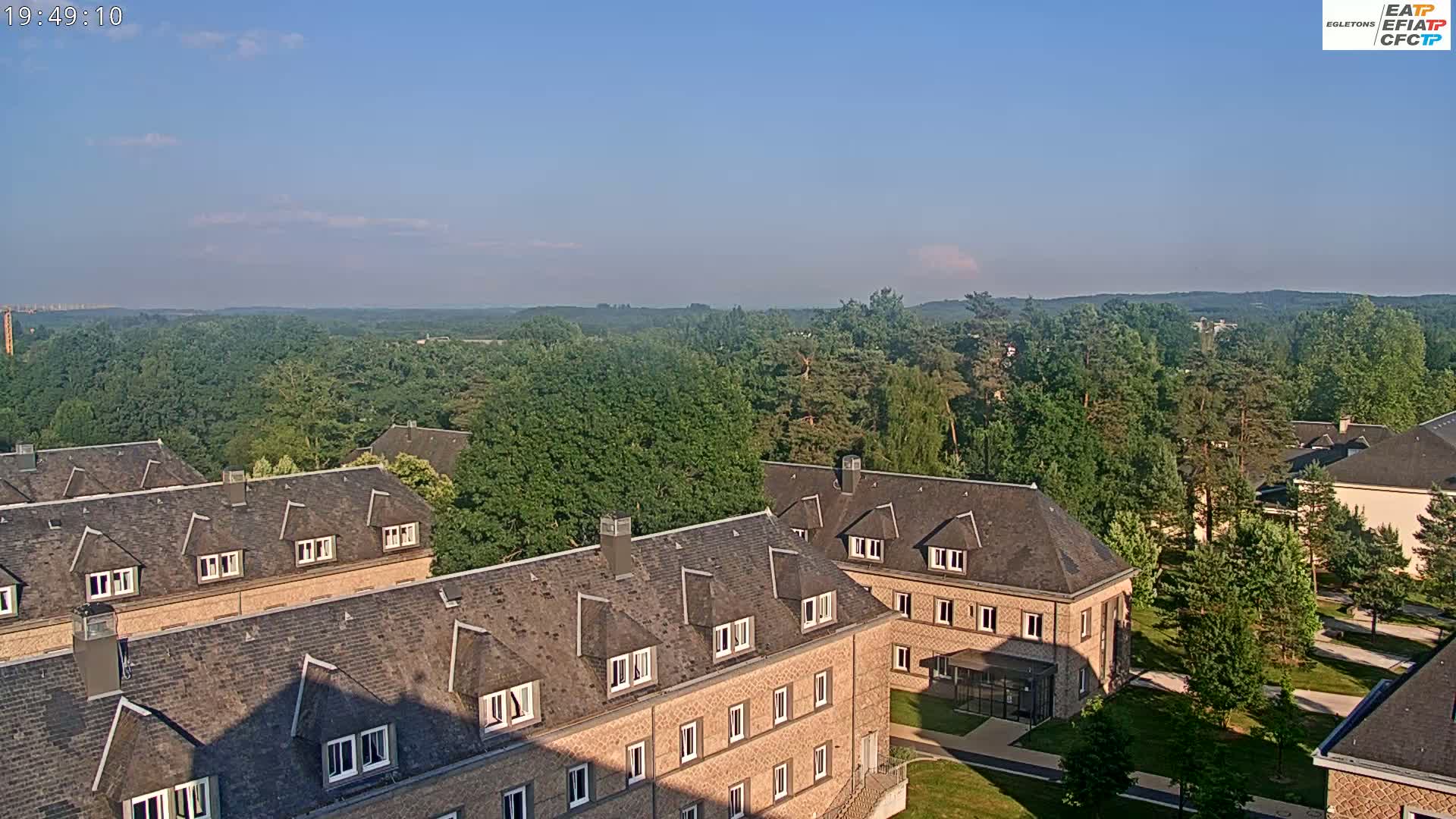 A high-angle view on a clear, sunny day shows several long, low brick buildings with dark gray roofs situated among green trees, with a large expanse of woods visible in the distance.