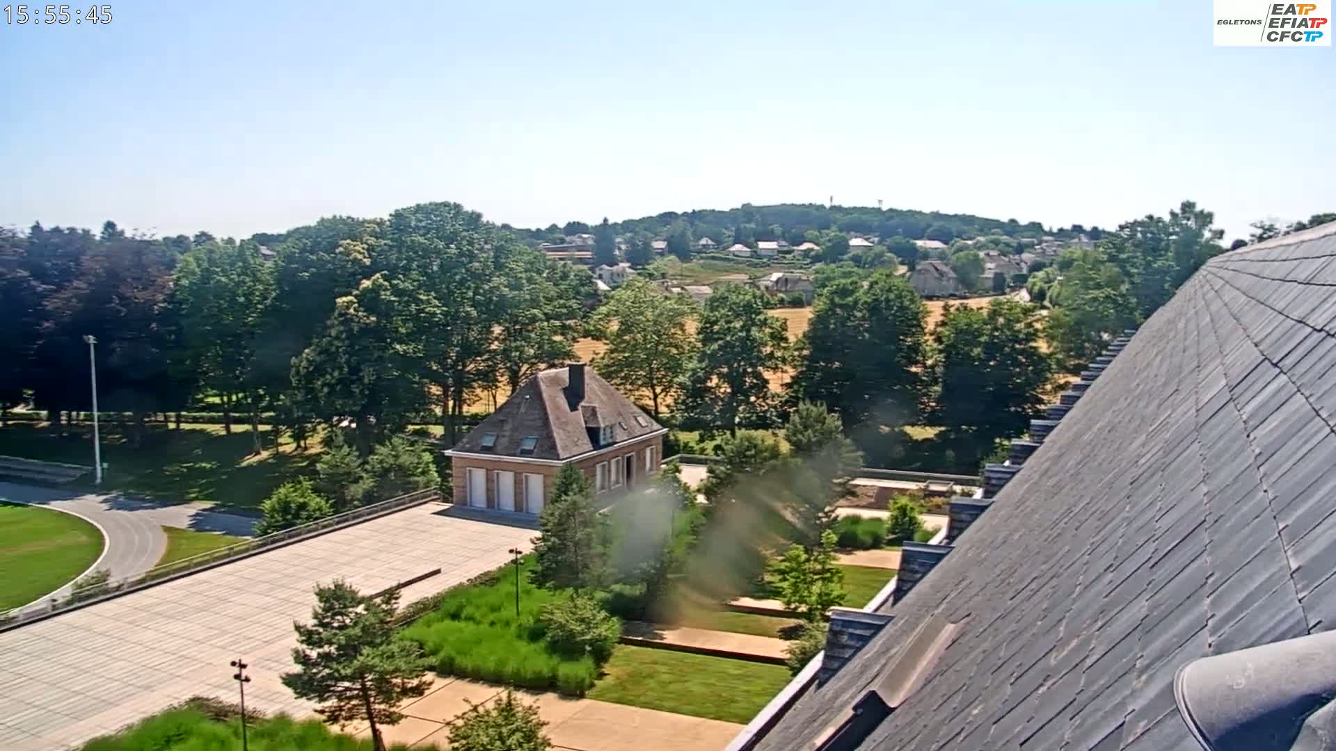 An elevated view of a landscaped courtyard with a small building, a running track, and a residential area in the background on a clear, sunny day.