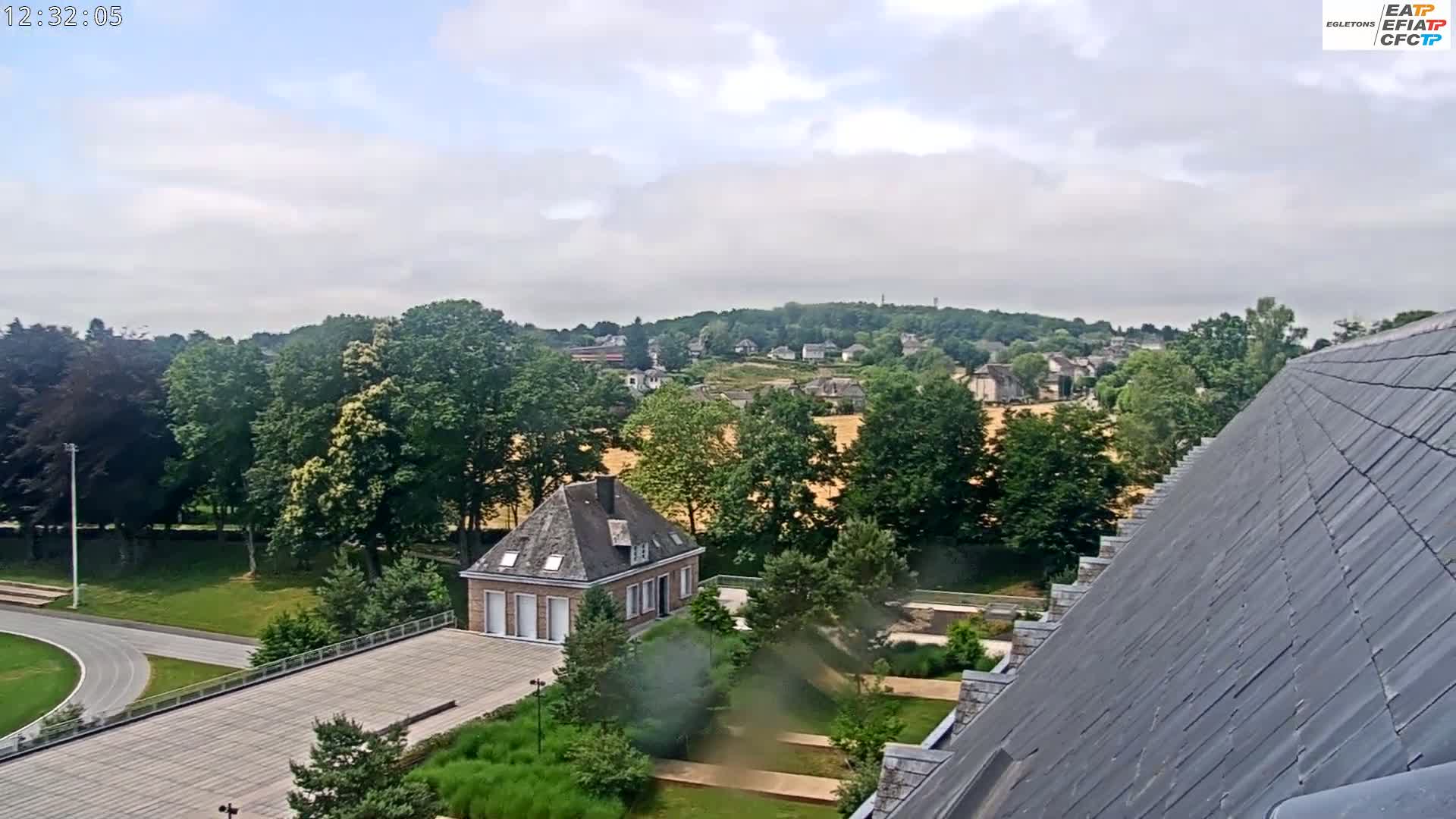 A partly cloudy day reveals a view of a small building, a running track, green lawns, and a residential area nestled among trees, with a portion of a sloped roof in the foreground.