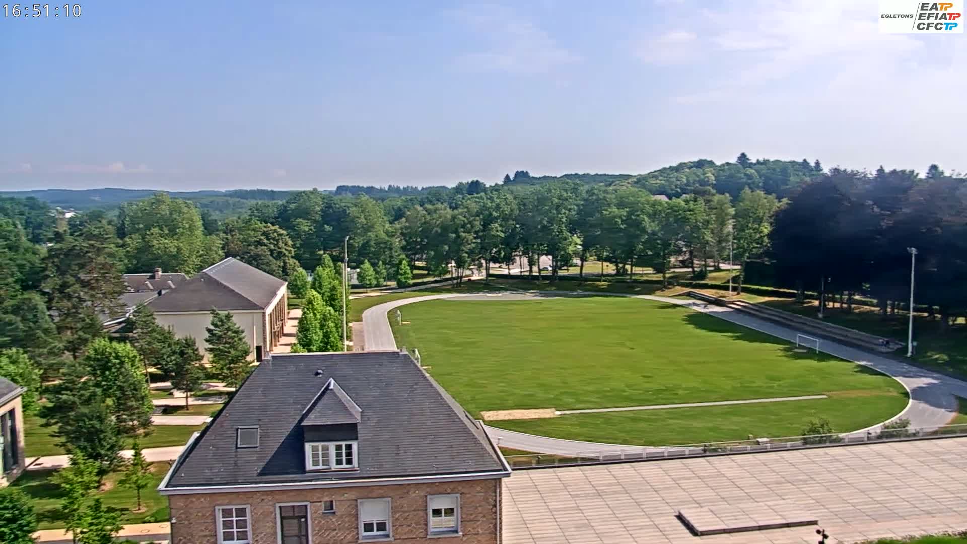 An aerial view shows a grassy athletic field with a running track, surrounded by buildings and lush green trees under a clear blue sky.