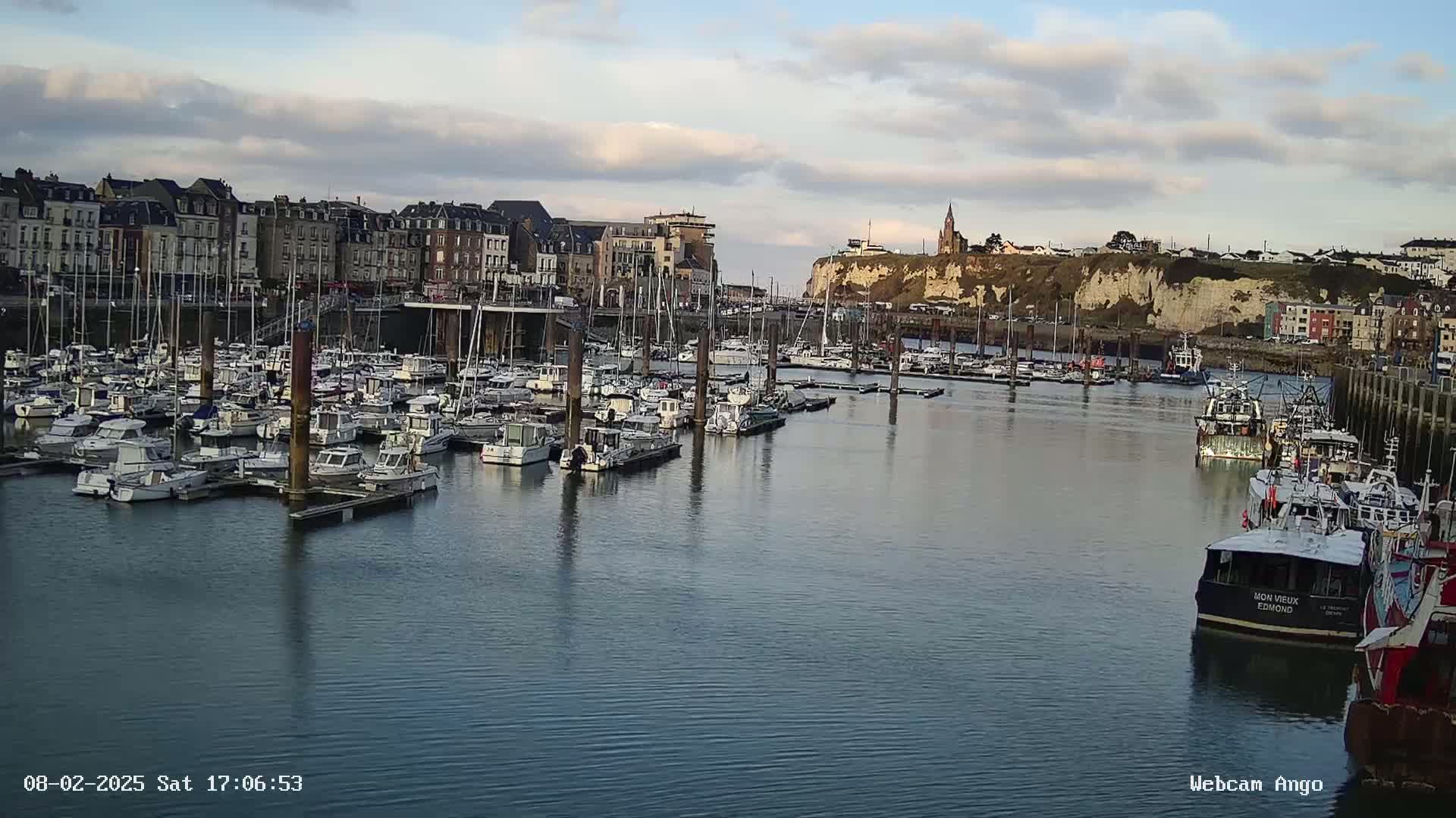 A harbor filled with numerous boats and yachts is situated in front of a town built against a cliffside, under a partly cloudy sky.