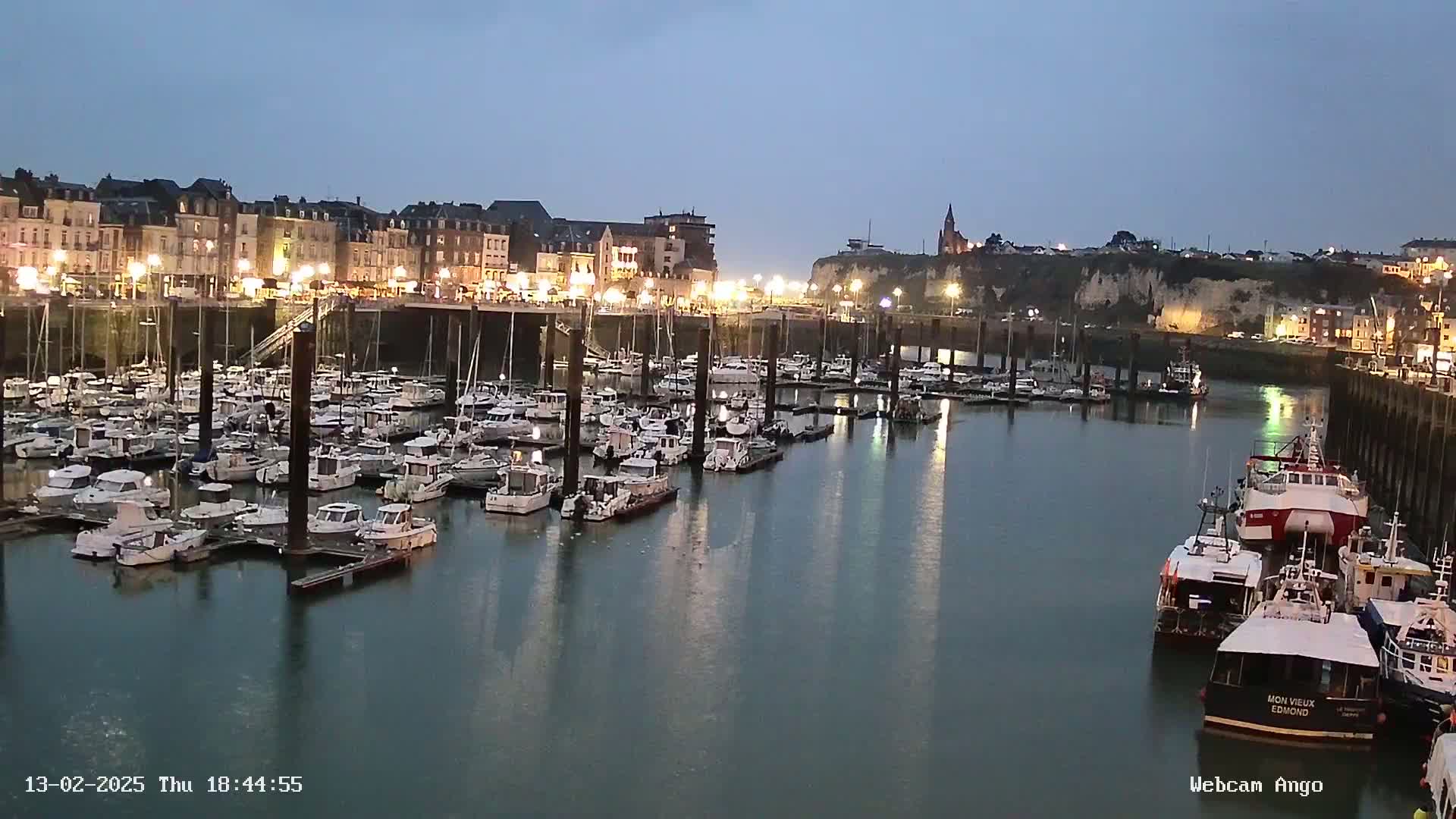 A harbor filled with many boats at night, under a twilight sky, is illuminated by lights along the waterfront buildings.