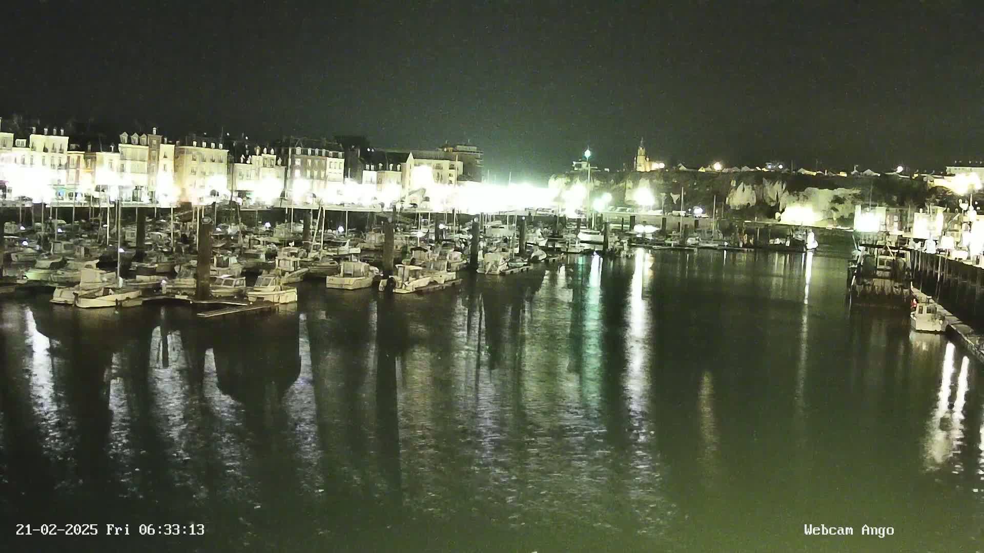 A nighttime view of a harbor filled with boats docked along a waterfront lined with buildings, reflecting in calm water.