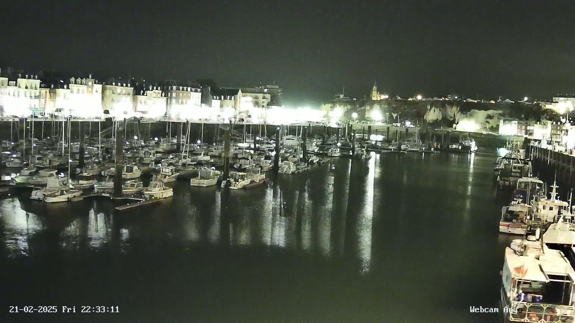 A nighttime view of a harbor filled with numerous boats docked along a waterfront lined with buildings, reflecting in the calm water.