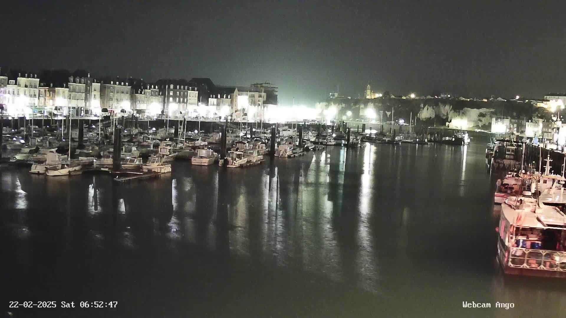 A nighttime harbor scene shows many boats moored at a dock, with buildings and cliffs illuminated in the background under a dark, possibly overcast sky.