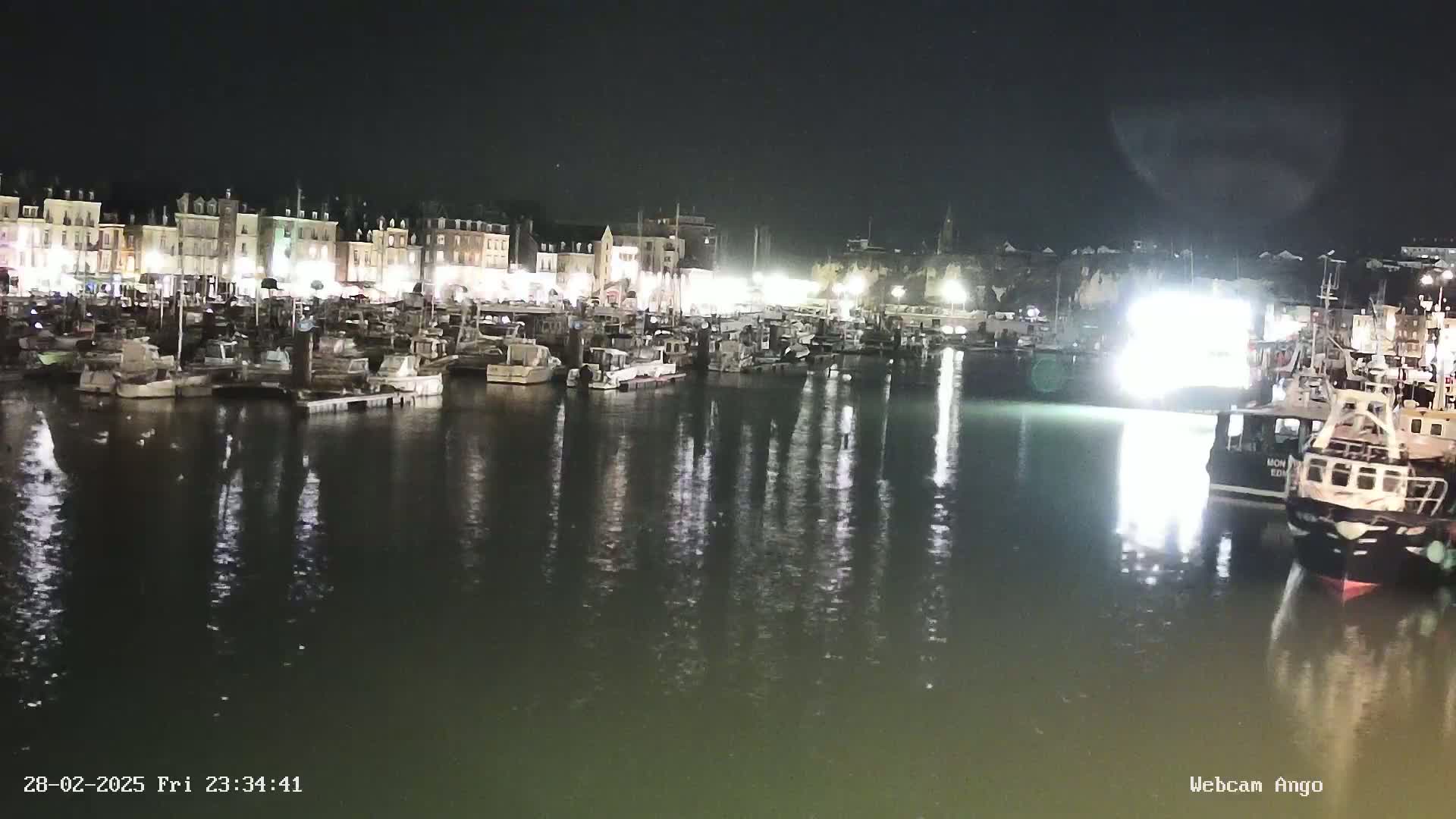A nighttime harbor scene shows many boats moored along a waterfront lined with buildings, all reflecting in calm water.