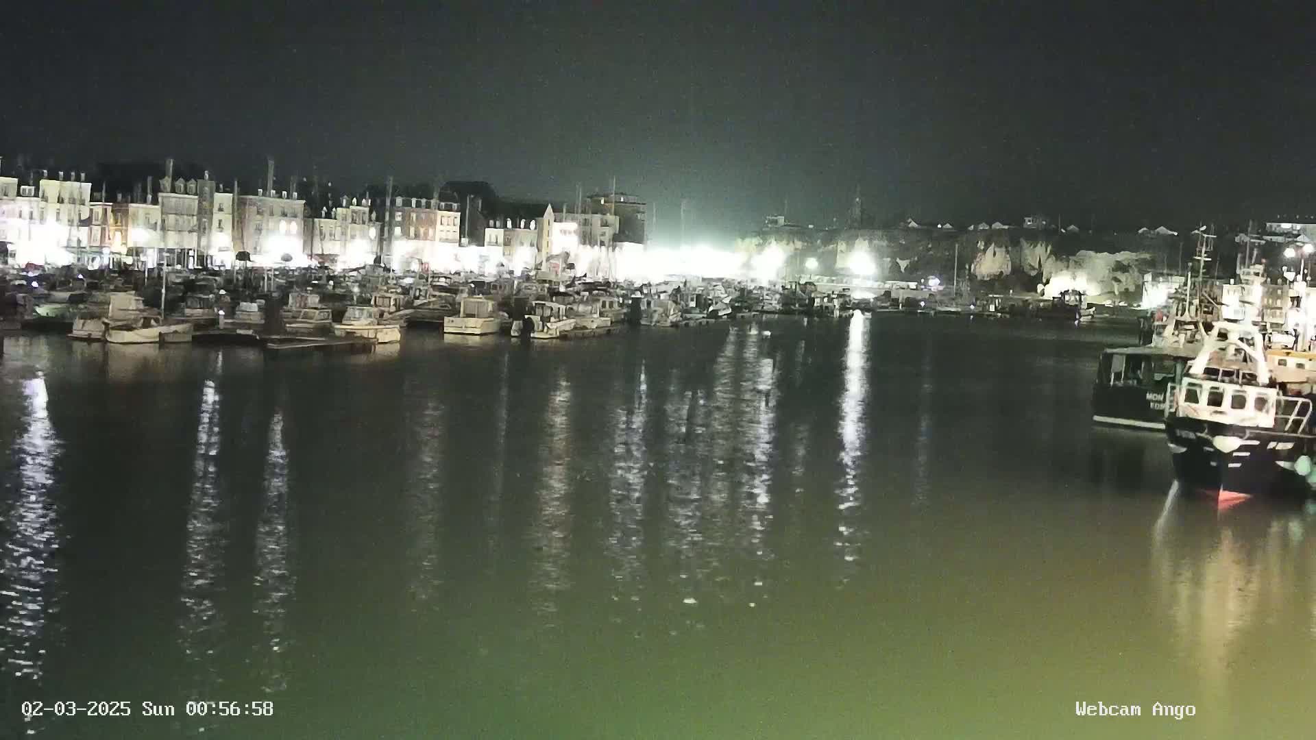 Many boats are moored in a harbor at night, illuminated by lights from buildings along the waterfront.