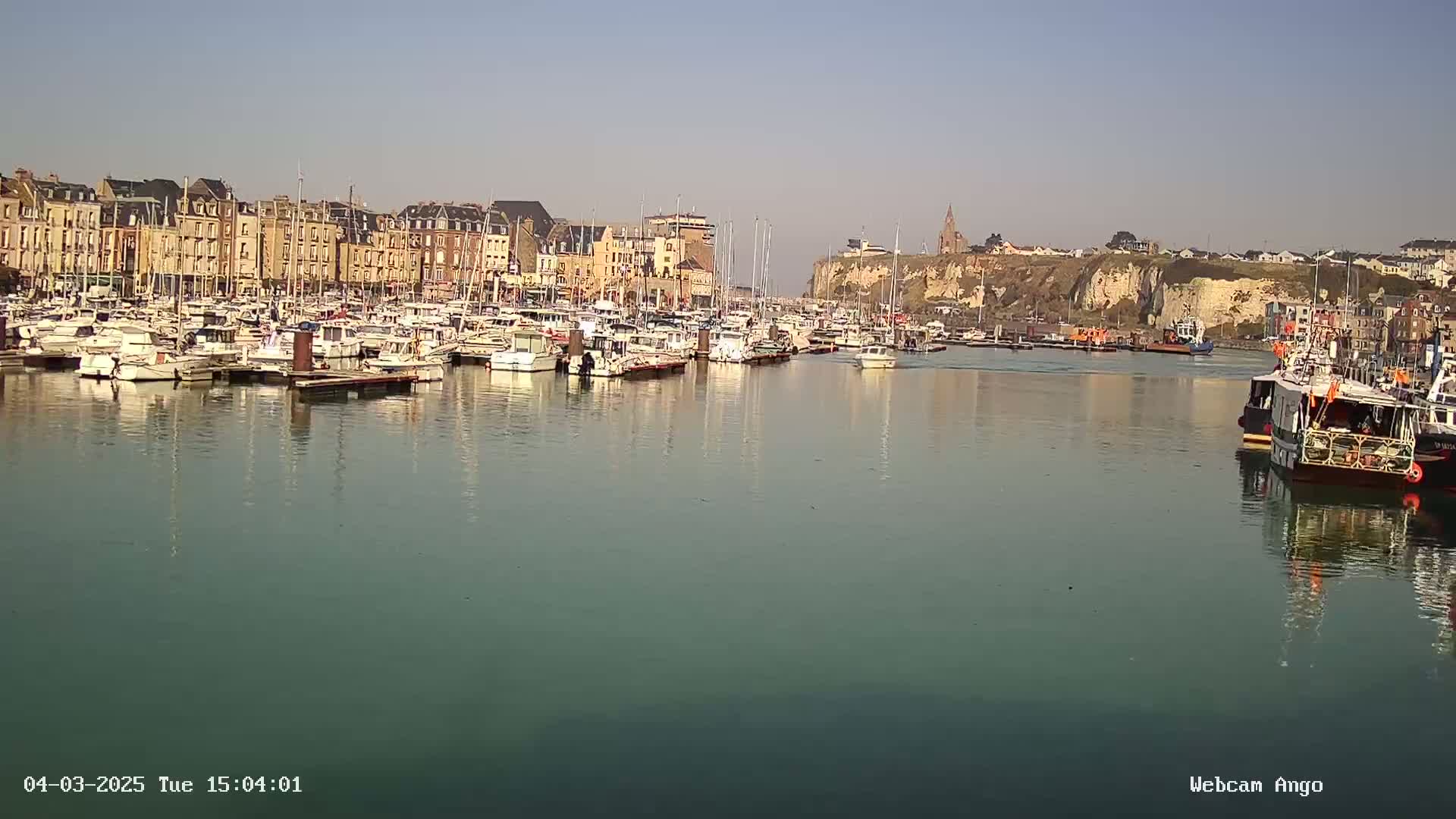 A calm harbor filled with numerous boats is situated in front of a town with multi-story buildings, a cliff, and a church, under a clear sky.