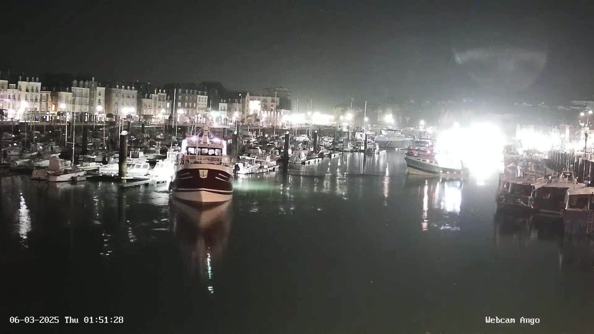 A nighttime harbor scene shows numerous boats docked along a waterfront lined with buildings, under a dark, possibly overcast sky.