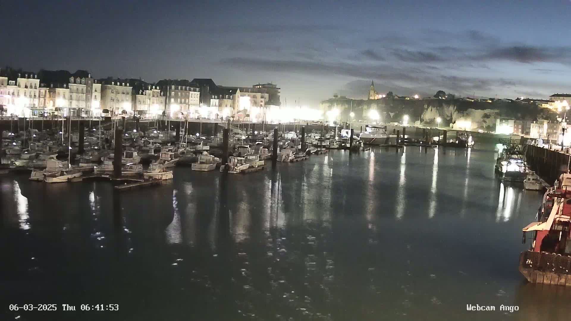 A nighttime harbor scene shows many boats docked along a waterfront lined with buildings under a partly cloudy sky.