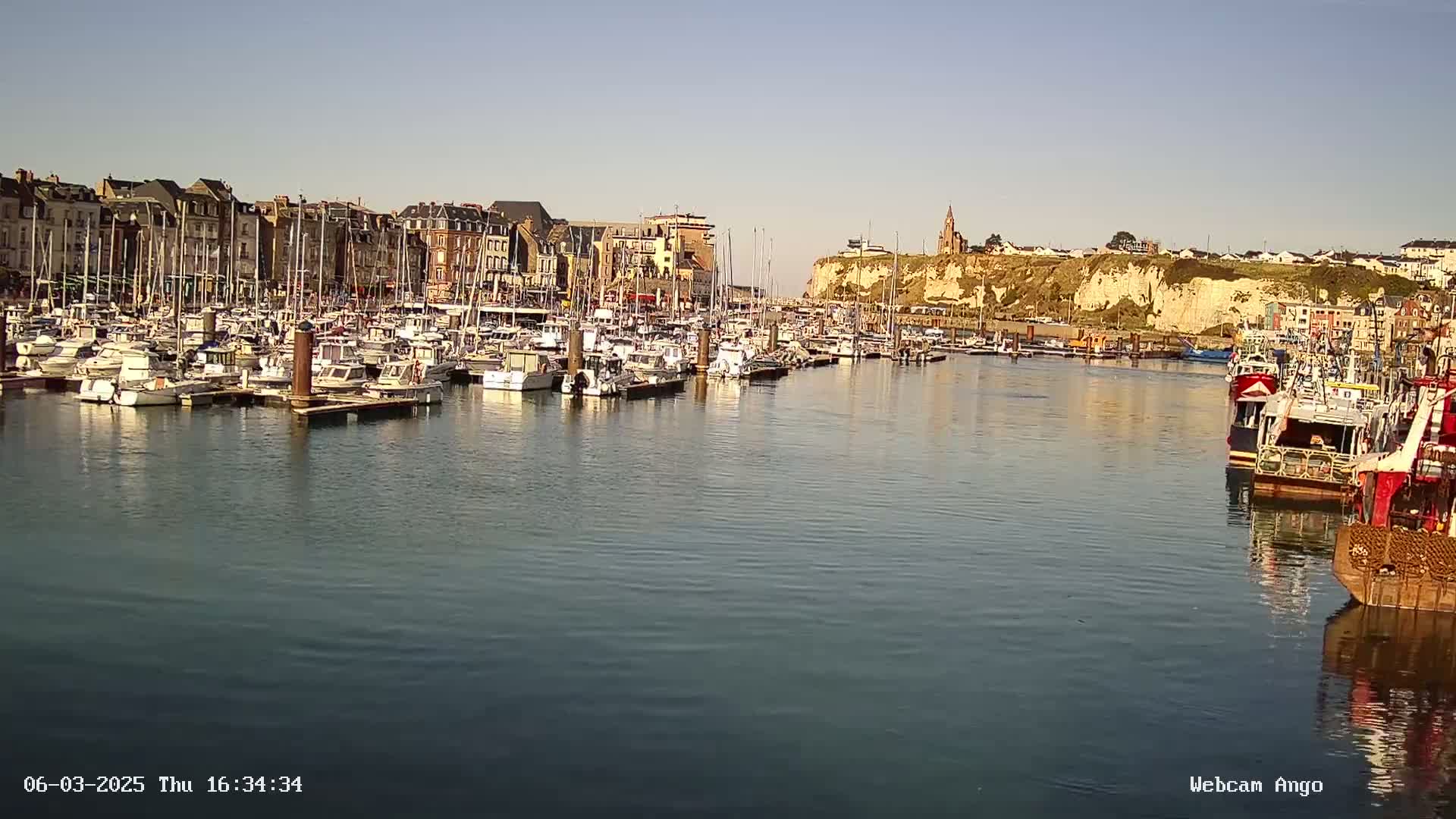 A calm harbor filled with numerous sailboats and fishing vessels is situated in front of a town with buildings lining the waterfront, backed by a tall cliff.