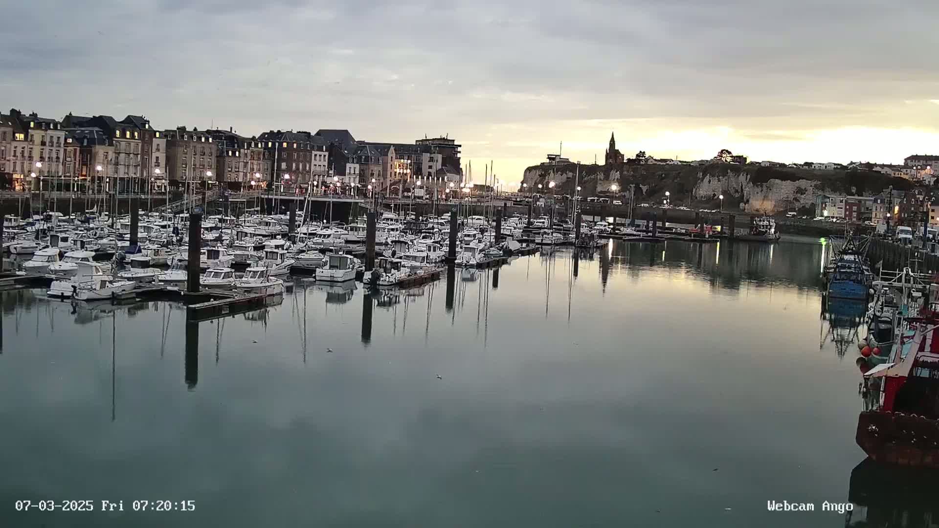 Many boats are docked in a harbor at twilight under a cloudy sky, with buildings and a cliff visible in the background.