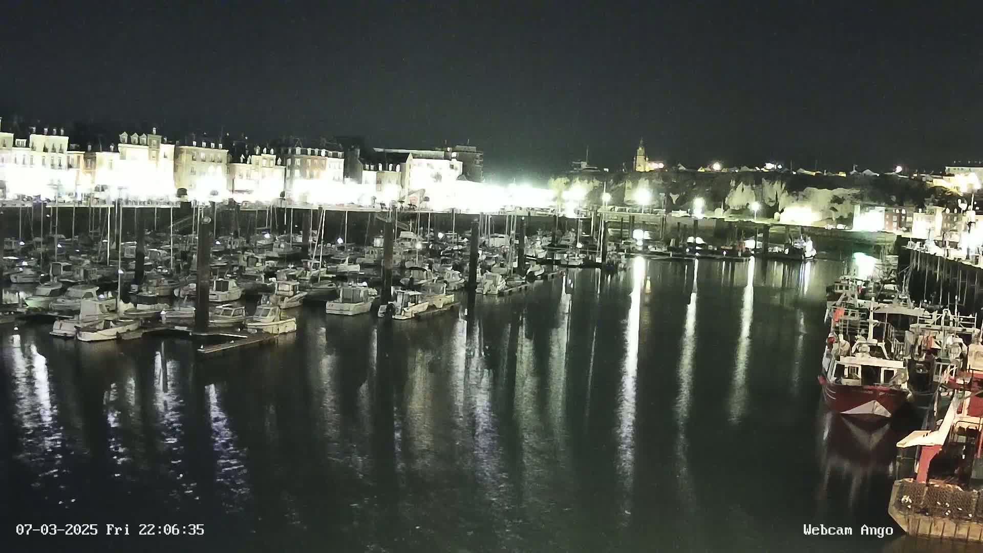 A nighttime view of a harbor filled with numerous boats docked alongside a waterfront lined with buildings, reflecting in the calm water.