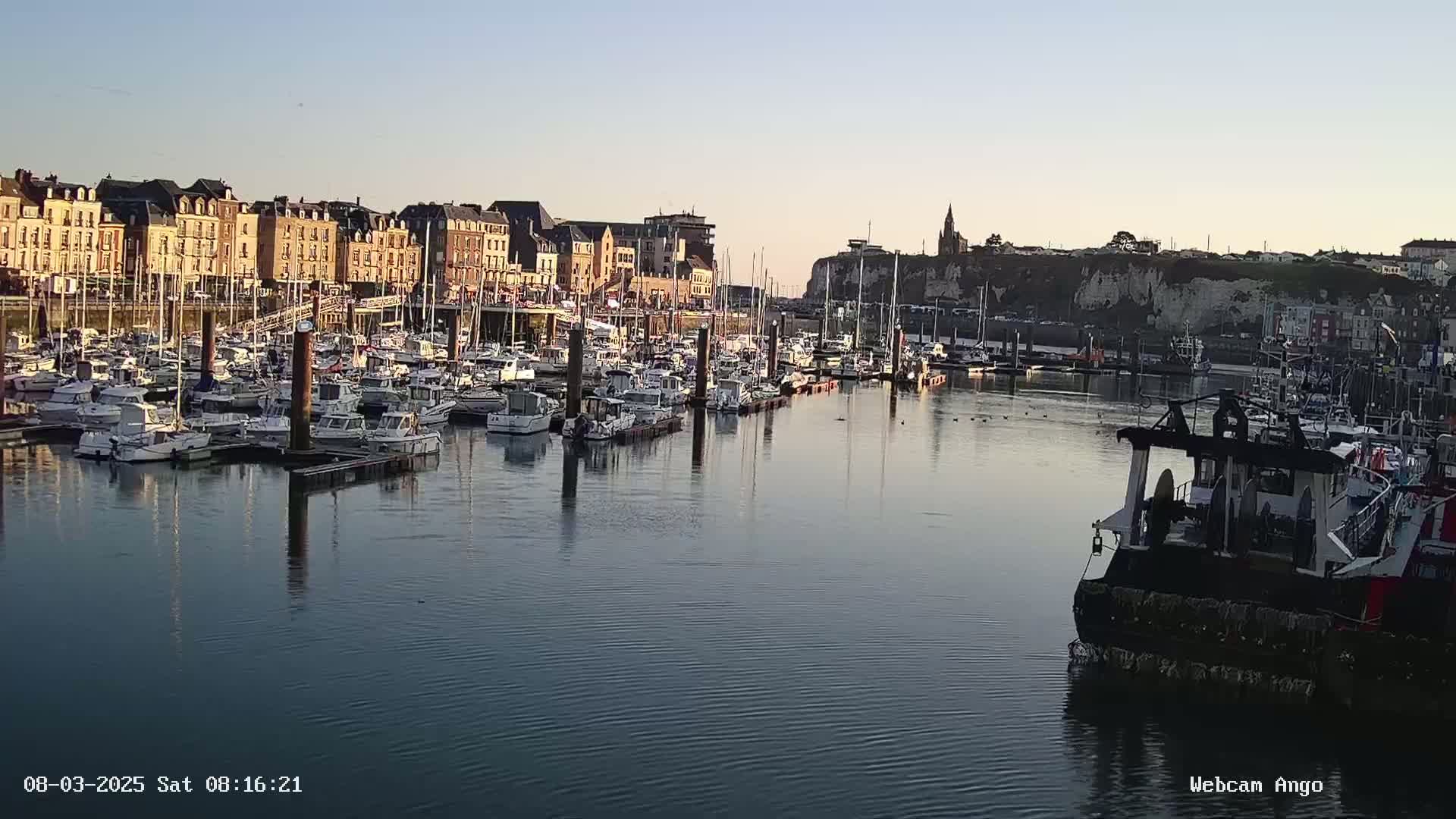 A calm harbor filled with numerous boats is situated in front of a waterfront town under a clear, twilight sky.