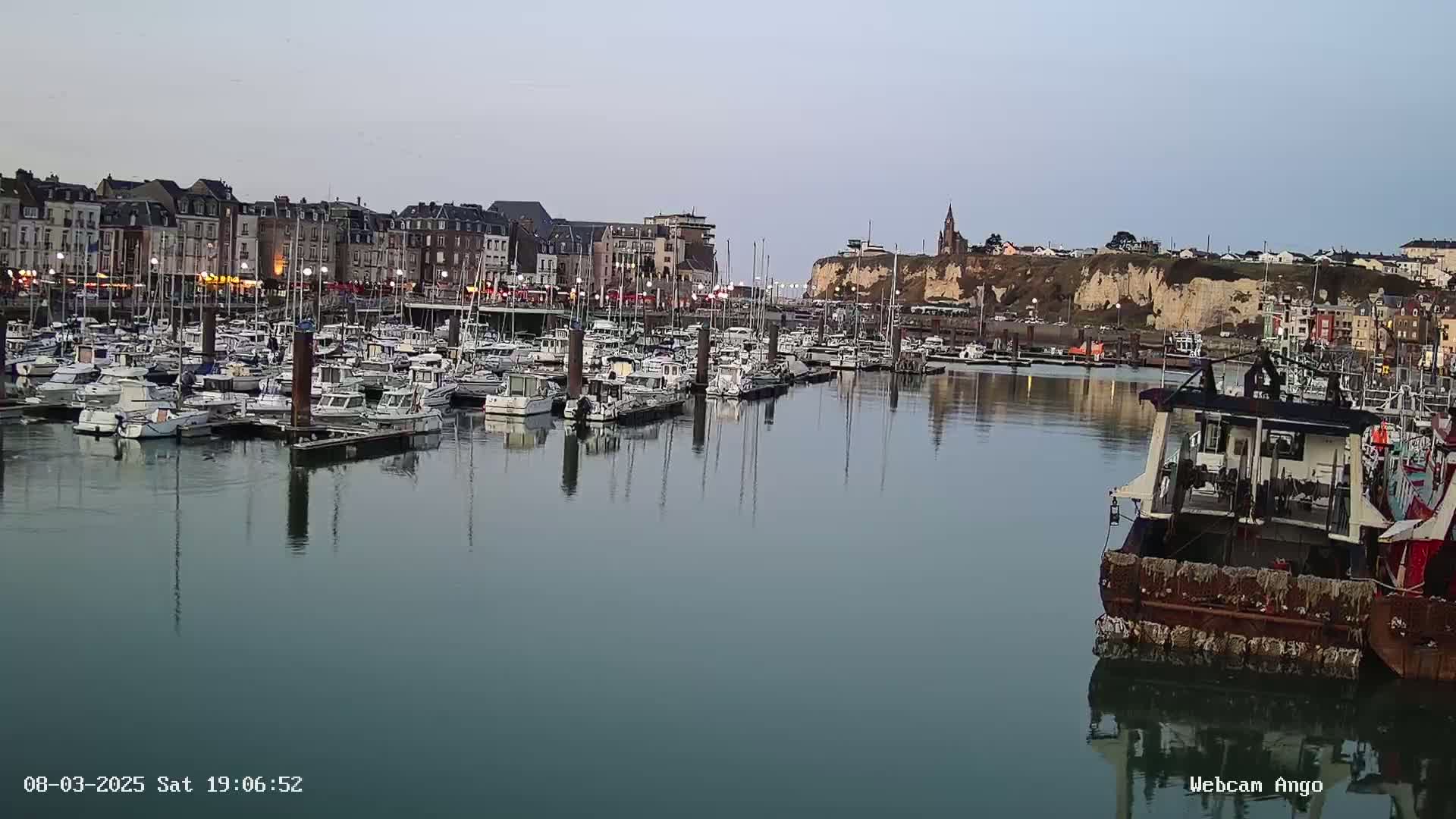 A calm harbor filled with numerous boats is situated in front of a town built along a cliff face under a dusky sky.
