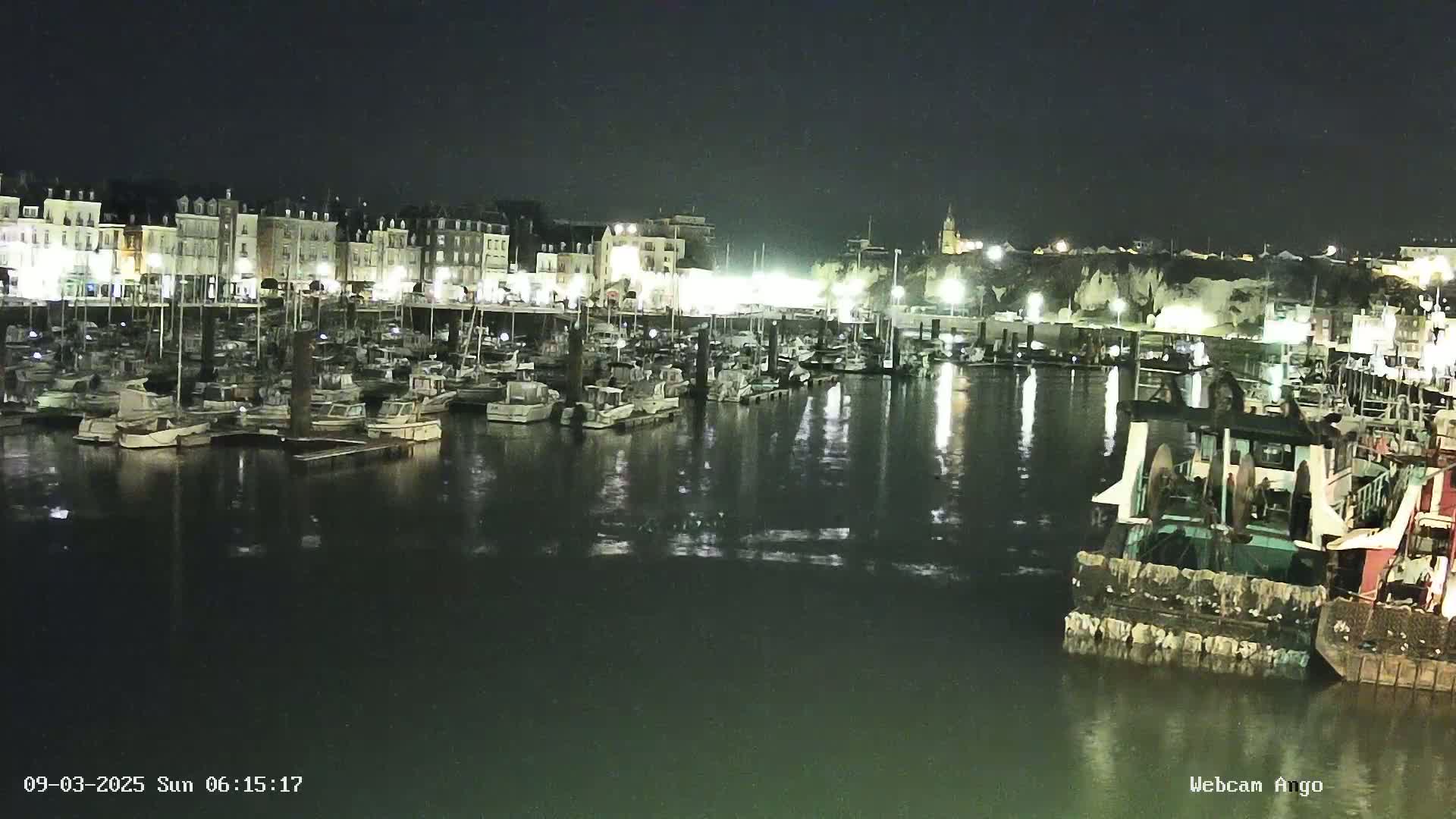 A nighttime harbor scene shows many boats moored at a dock in front of brightly lit buildings, under a clear, dark sky.