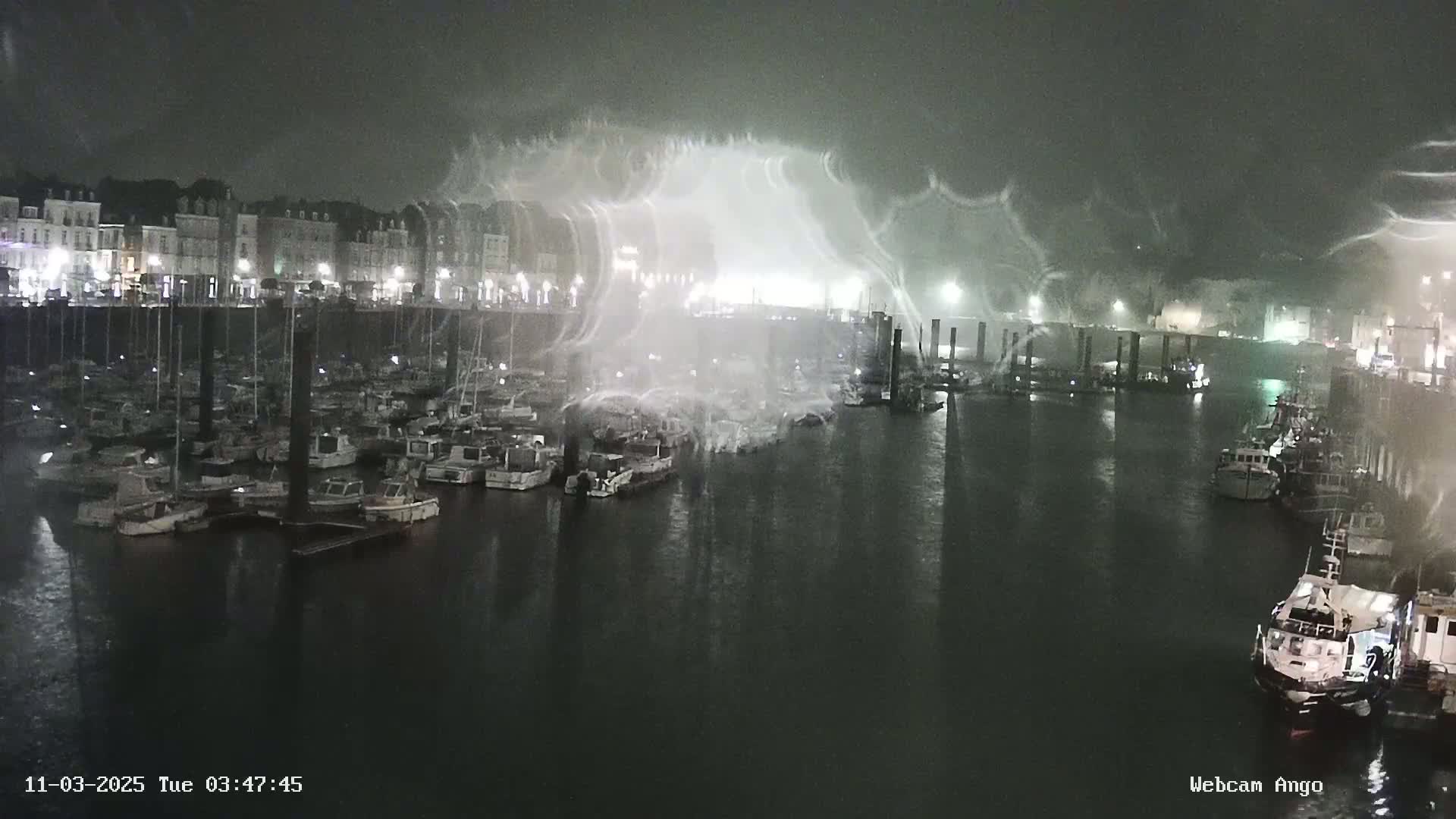 A nighttime harbor filled with boats is shown under heavy, spray-like rain or mist.