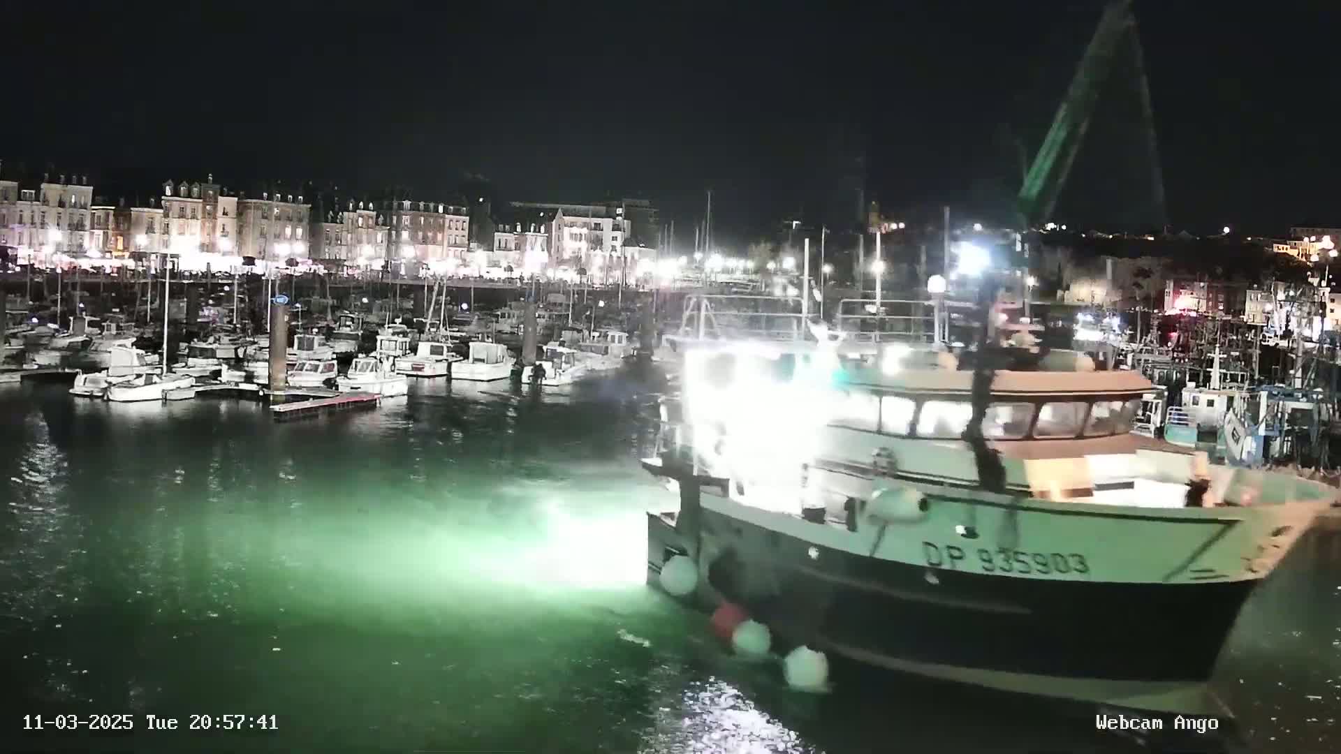 A large fishing boat is maneuvering in a harbor at night, with many smaller boats moored along a waterfront lined with buildings.