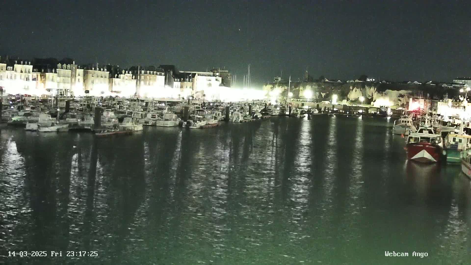 A nighttime harbor scene shows many boats moored at a dock, illuminated by lights from buildings along the shore.