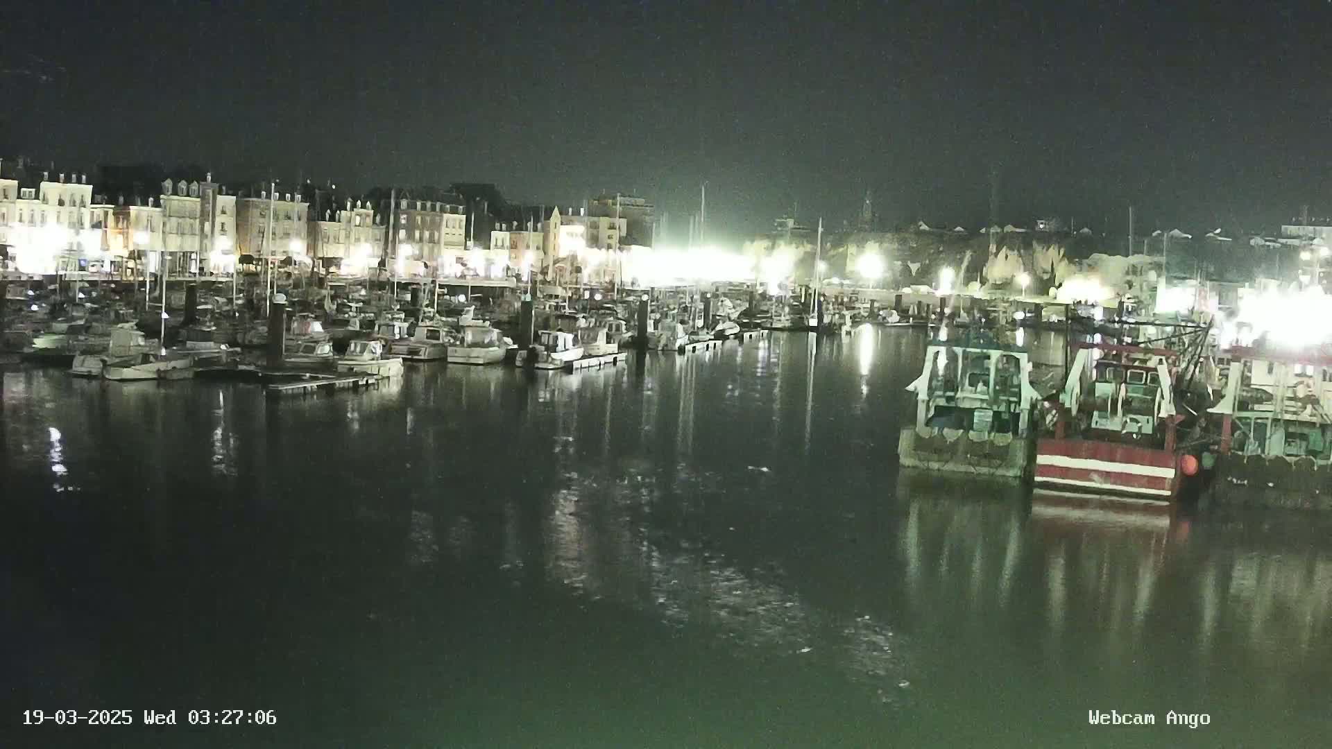 A nighttime harbor scene shows many boats moored in calm water, illuminated by lights from buildings along the waterfront.