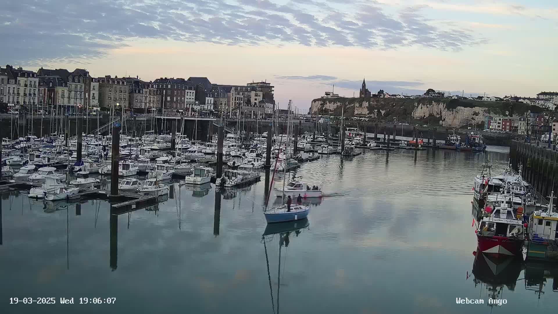A calm harbor filled with numerous sailboats and motorboats is situated in front of a town with multi-story buildings under a partly cloudy sky.