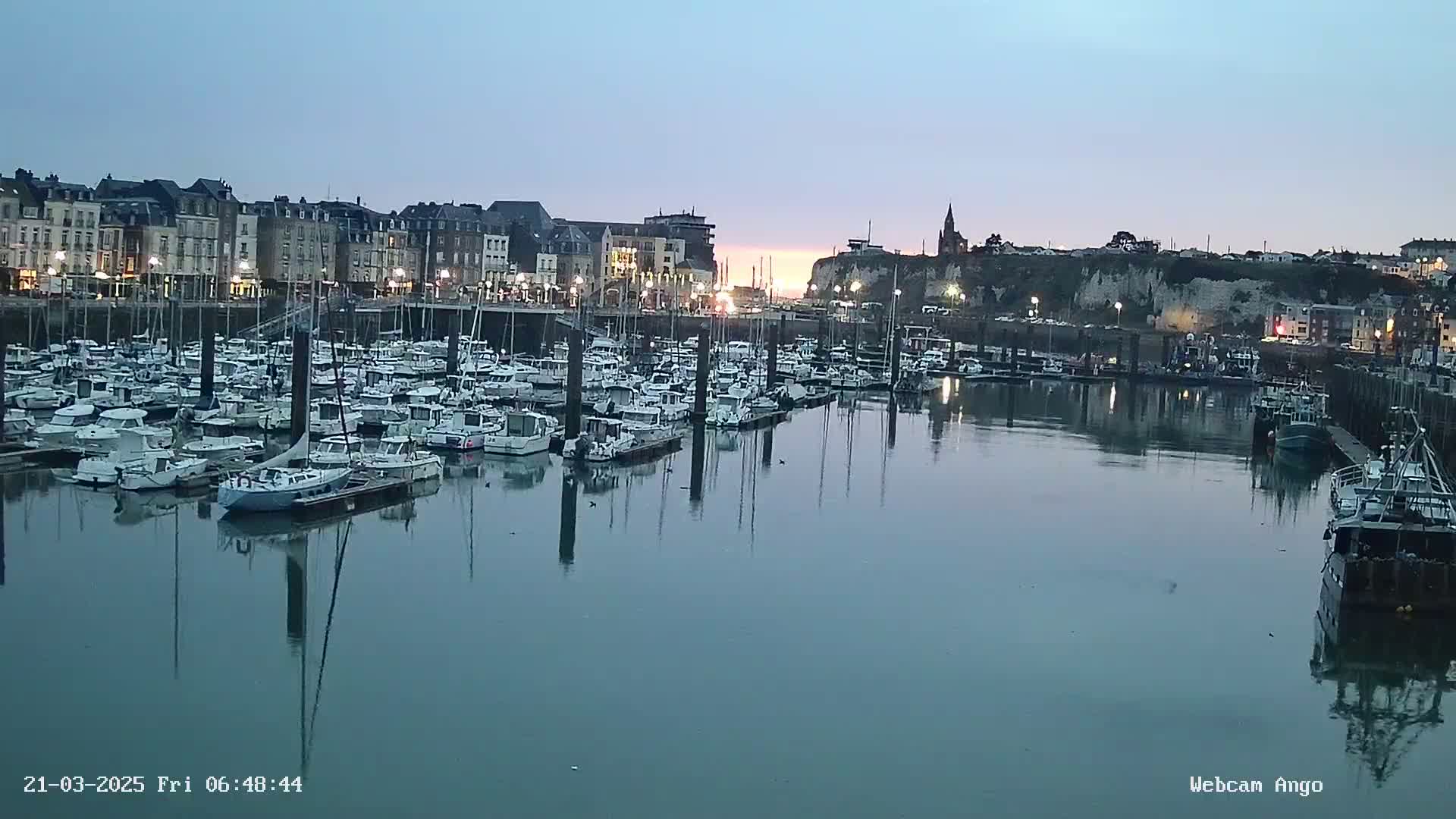 A calm harbor filled with numerous docked sailboats and motorboats at twilight, with a town and cliff in the background.