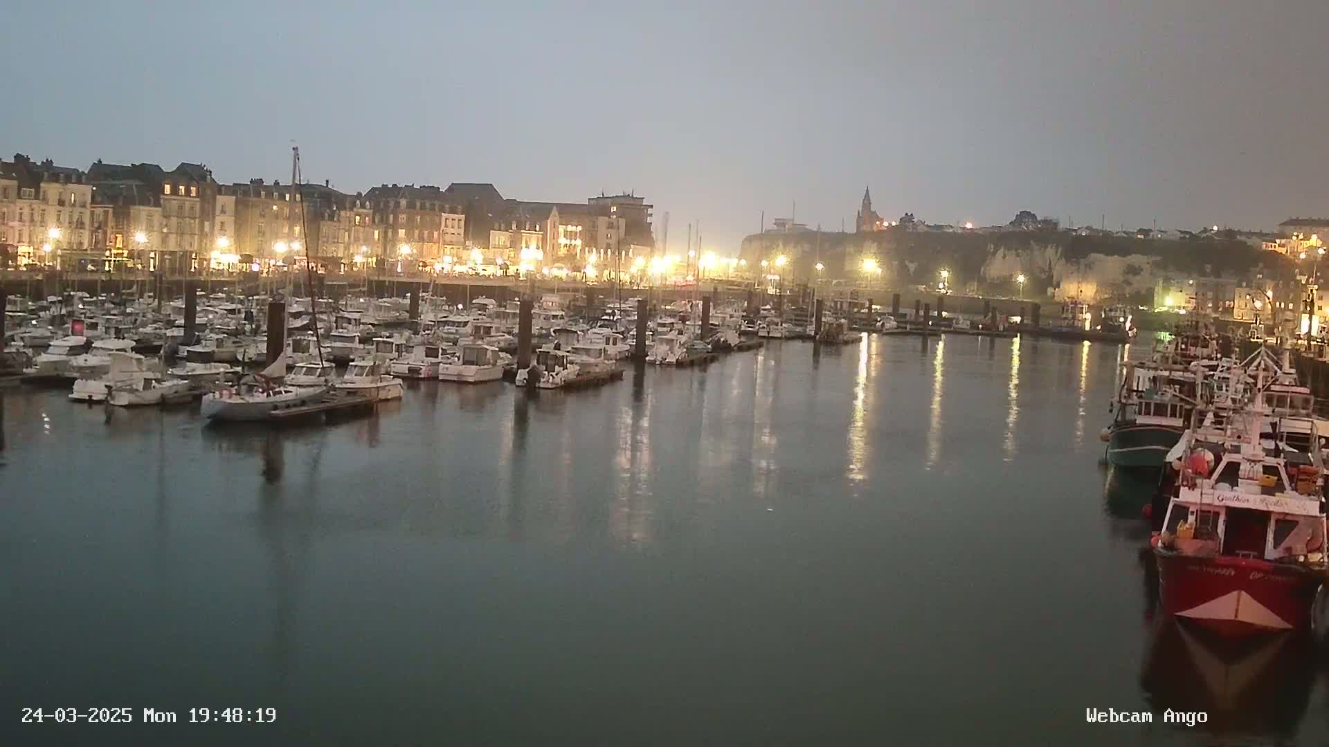 Many boats are docked in a harbor at dusk under an overcast sky, with a town's lights reflecting on the water.