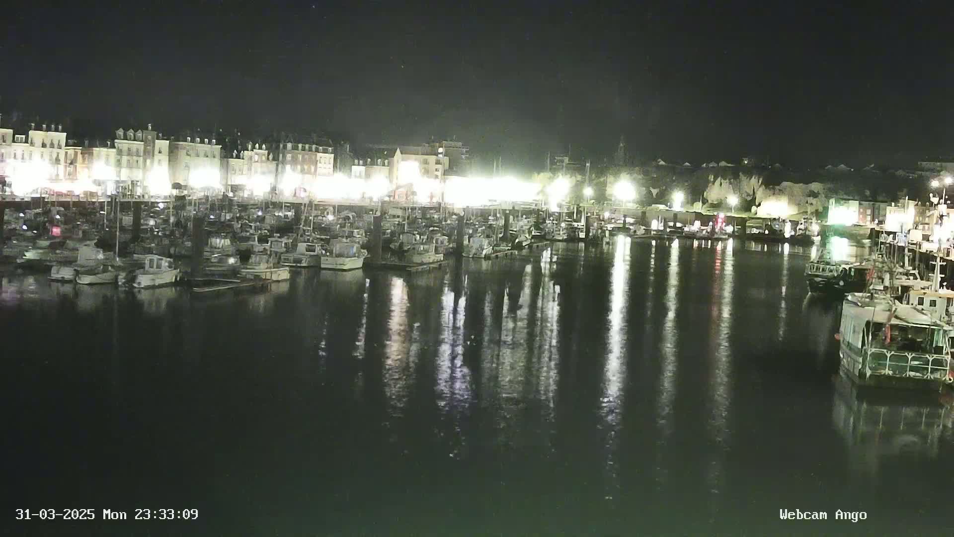 A nighttime harbor scene shows many boats moored along a waterfront lined with buildings, their lights reflecting on the calm water.