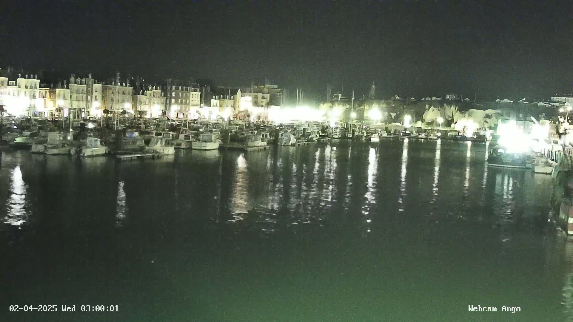 A nighttime harbor scene shows many boats moored along a waterfront lined with illuminated buildings.