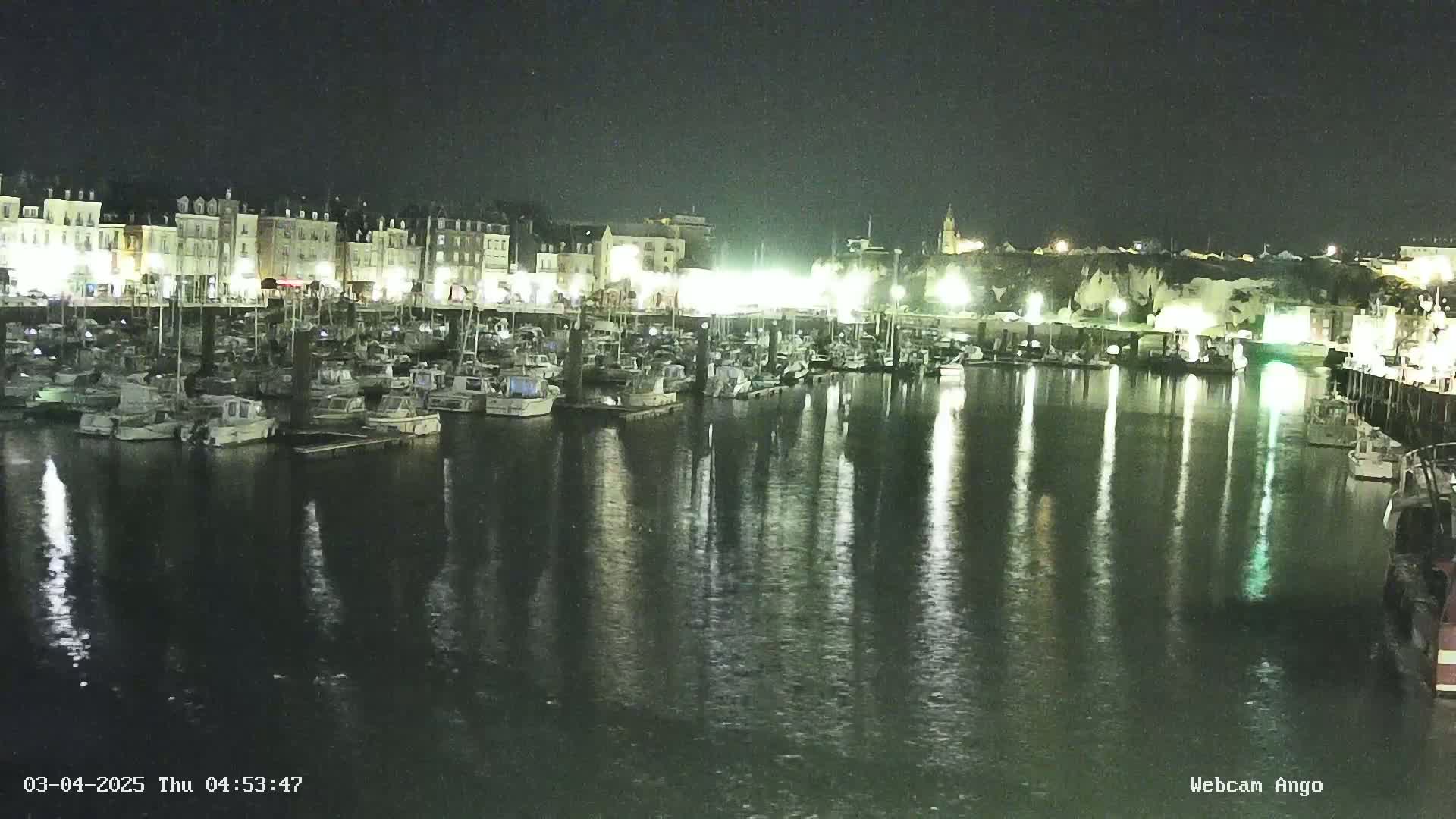 A nighttime view of a harbor filled with boats, reflecting the lights of buildings along the waterfront.