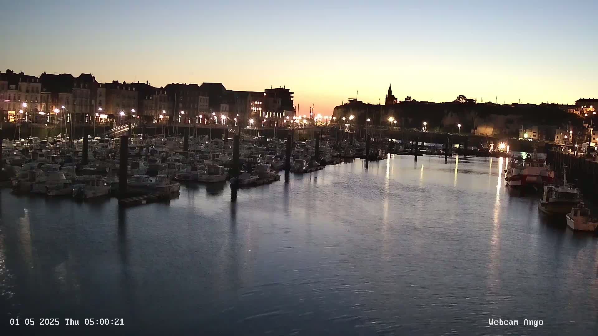 Many boats are moored in a harbor at twilight, with a town's buildings and lights visible in the background.