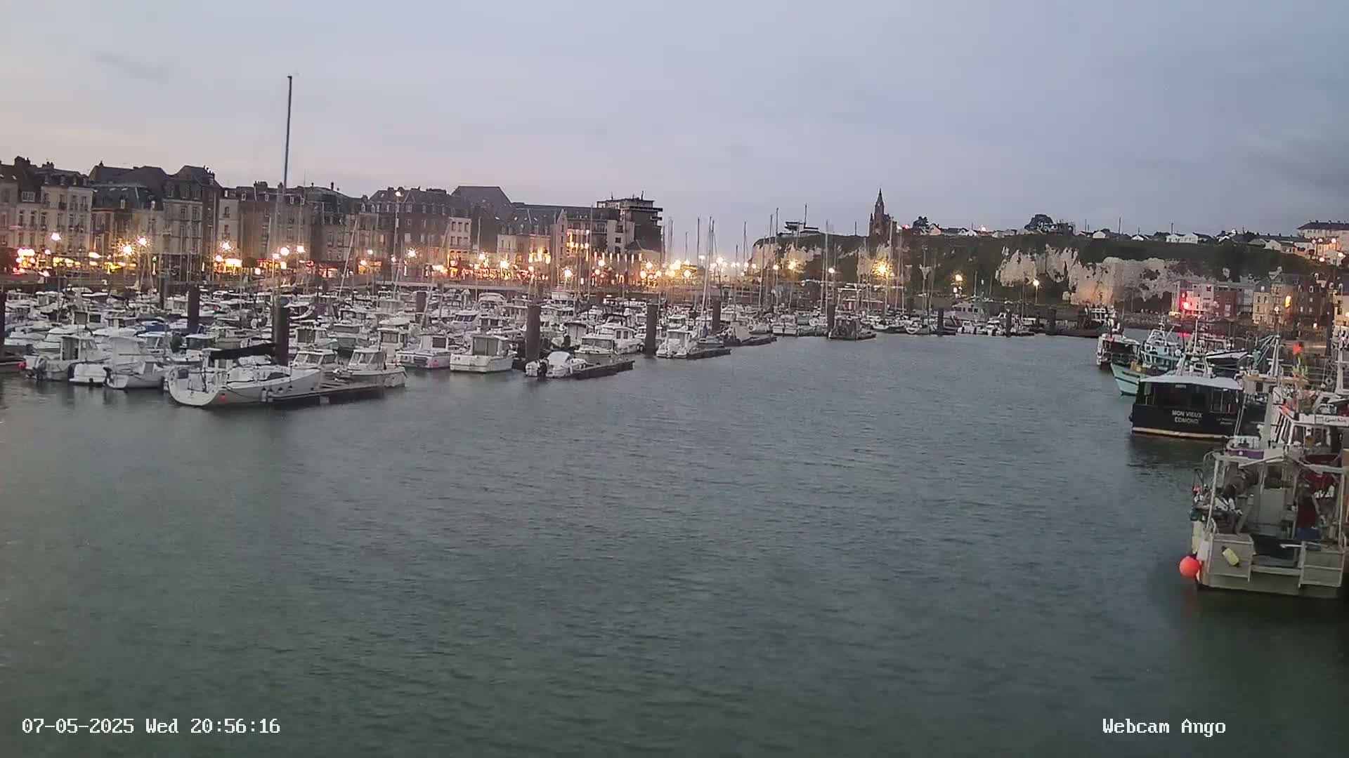 Many boats are docked in a harbor at dusk, under a mostly clear sky, with a town and cliffs visible in the background.