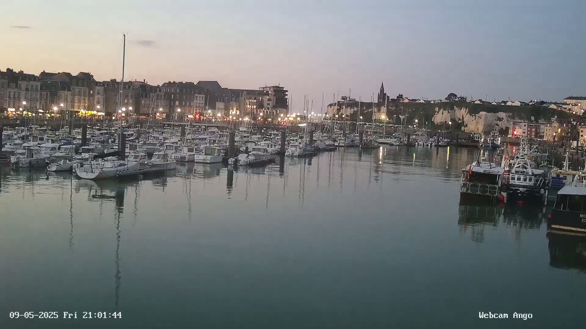A harbor filled with numerous boats is situated at dusk next to a town with buildings and cliffs visible in the background.