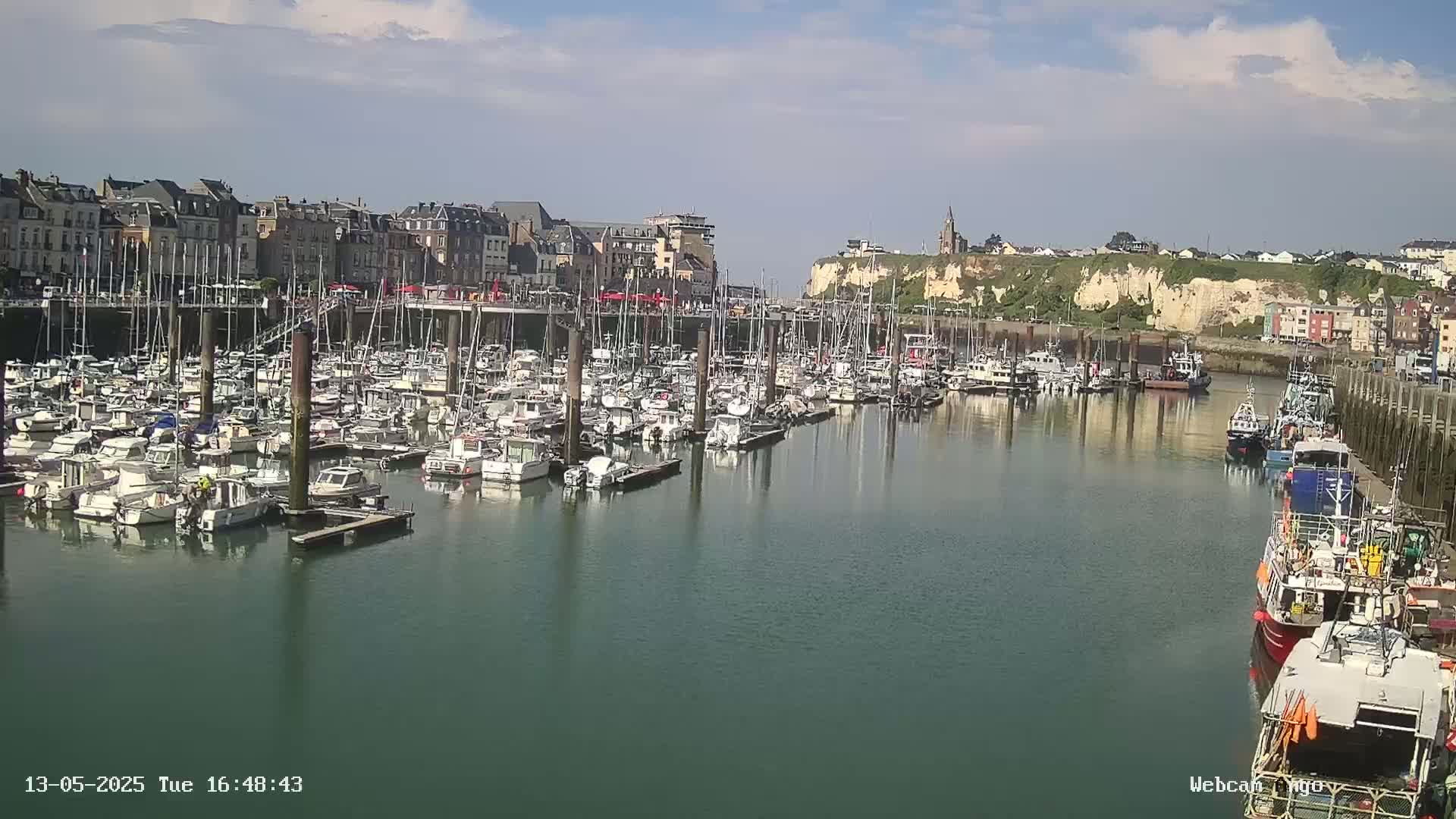 A harbor filled with numerous boats and yachts is nestled beside a town under a partly cloudy sky.