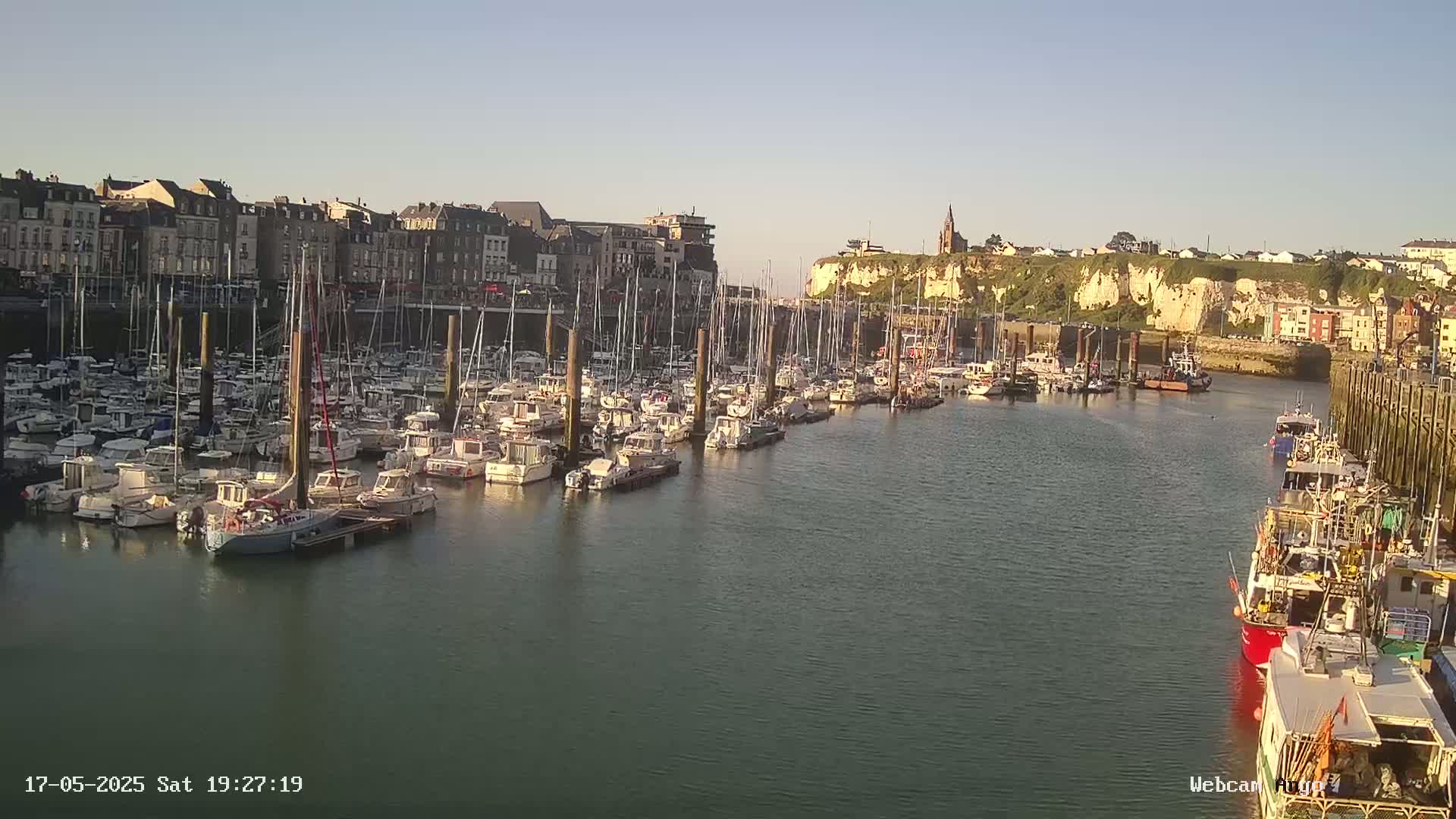 A calm harbor filled with numerous boats is nestled beside a town built along a cliffside on a clear day.