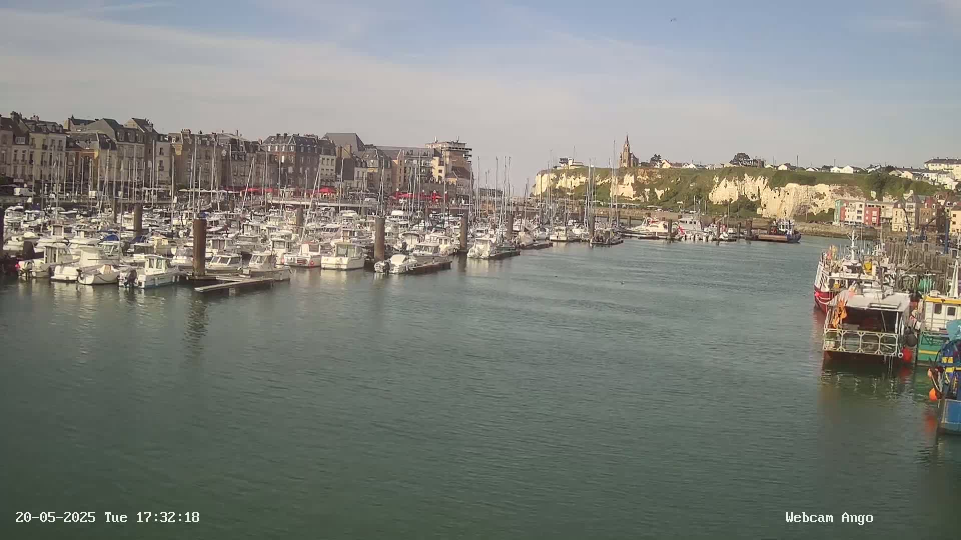 A harbor filled with many boats is nestled next to a town with buildings lining the waterfront, under a mostly sunny sky.