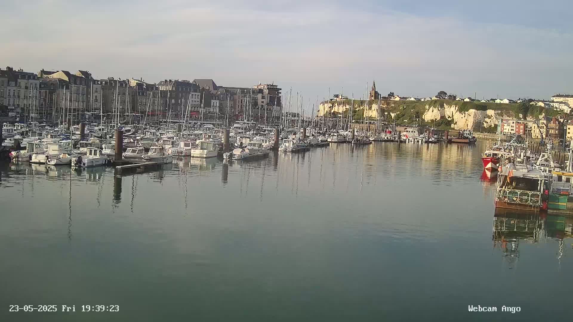 A calm harbor filled with many boats is nestled beside a town built against a high, white cliff face under a mostly clear sky.