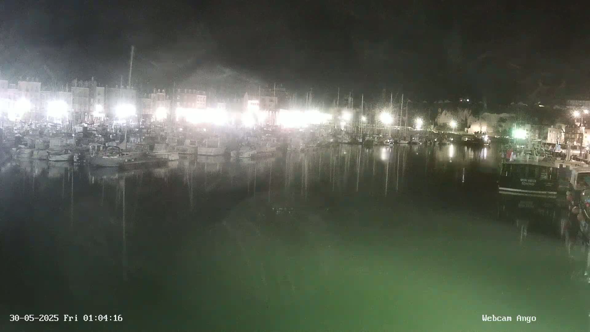 A nighttime harbor scene shows many boats moored at a dock, their lights reflecting in the dark, calm water.