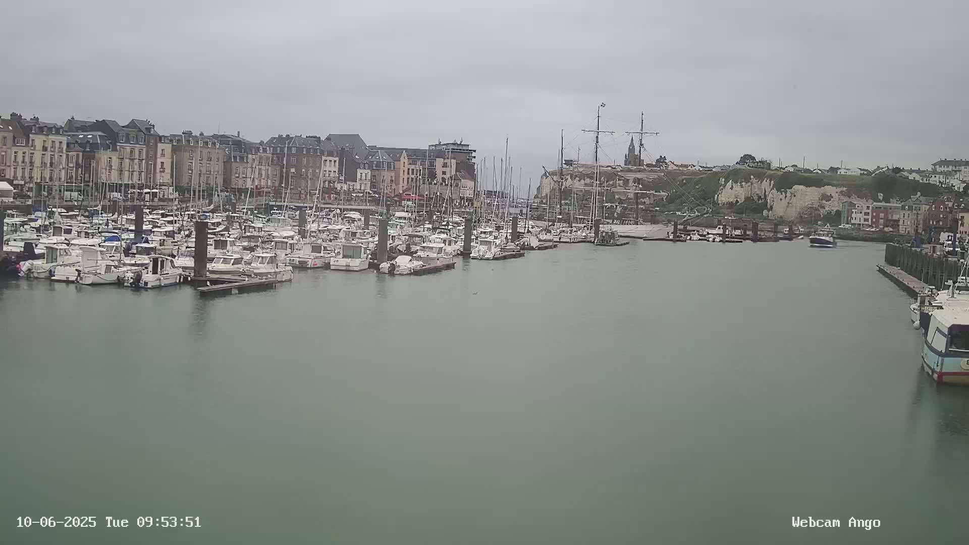 Many boats are moored in a harbor on an overcast day, with buildings lining the waterfront and white cliffs visible in the background.