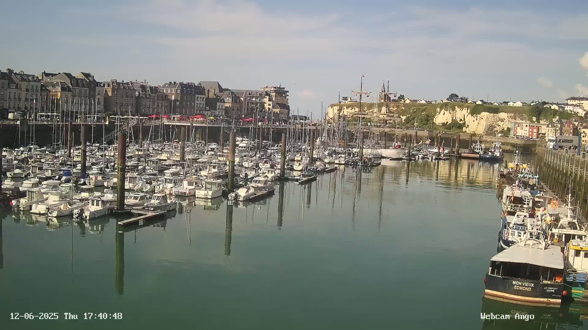 Many boats are docked in a harbor next to a town situated on a cliffside under a partly sunny sky.