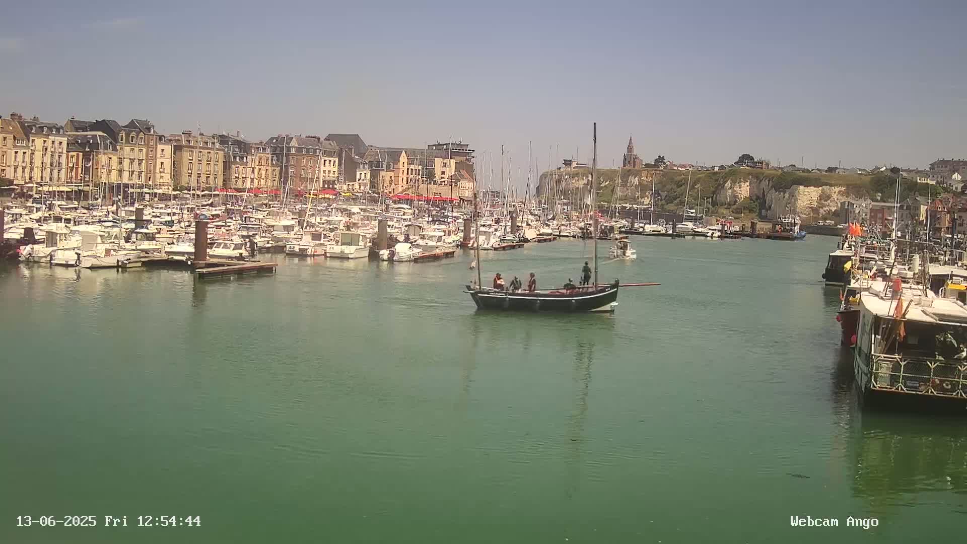 A harbor filled with numerous boats, a few of which are sailing, is situated in front of a town with multi-story buildings under a clear, sunny sky.