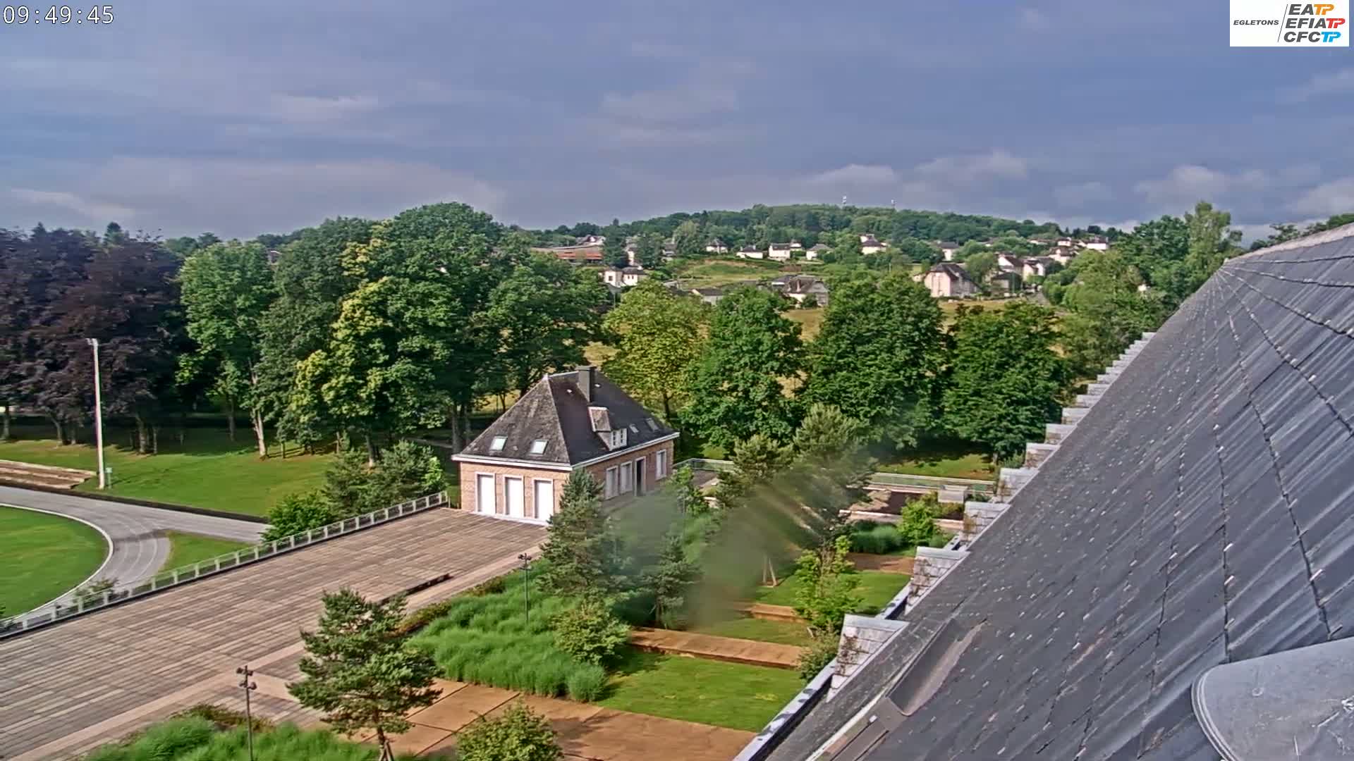A partly cloudy day overlooks a park with a small brick building and a running track, situated near a larger building with a sloped slate roof and a residential area in the background.