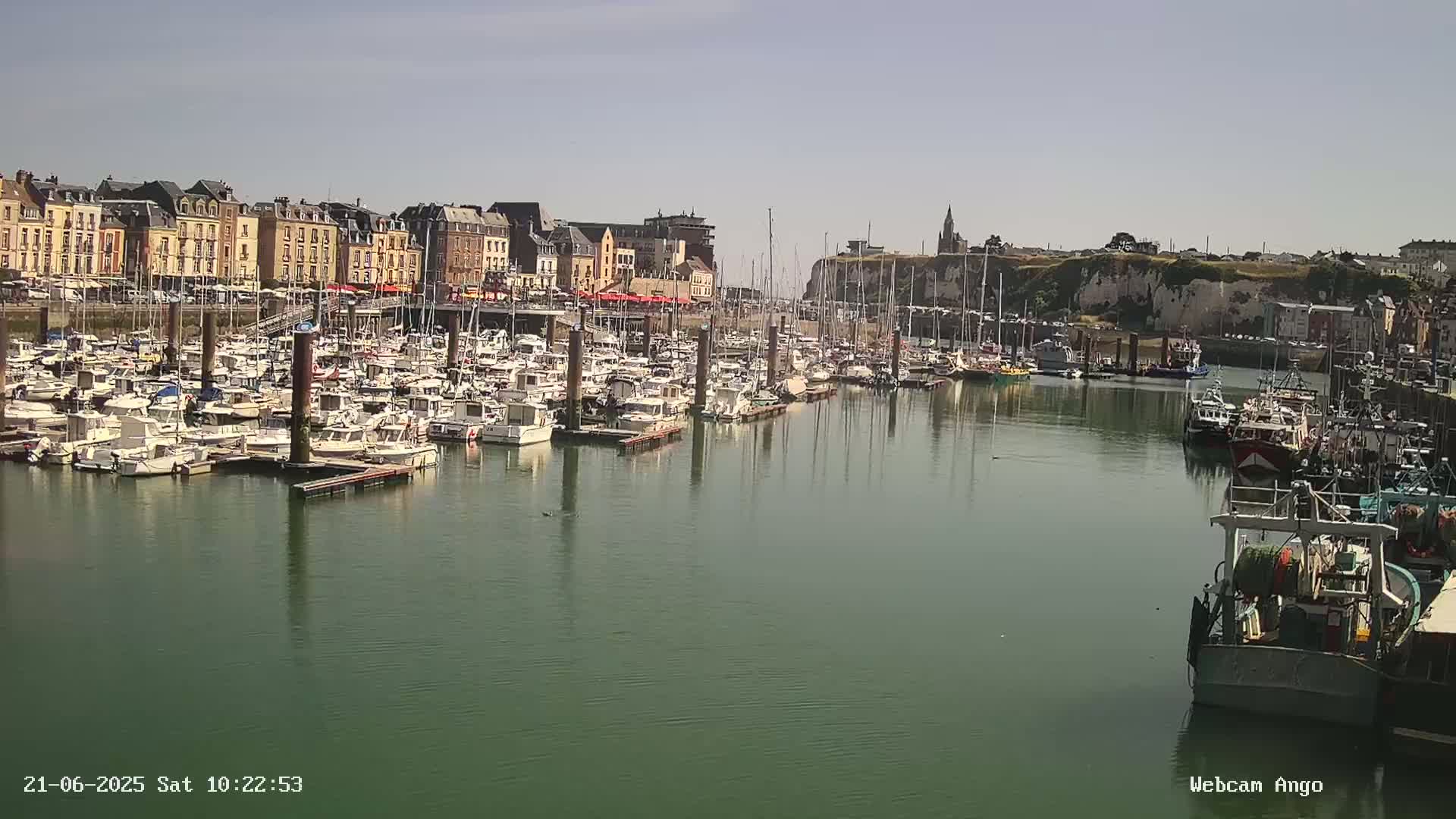 A calm harbor filled with numerous docked boats and yachts sits before a town of multi-story buildings under a sunny sky.