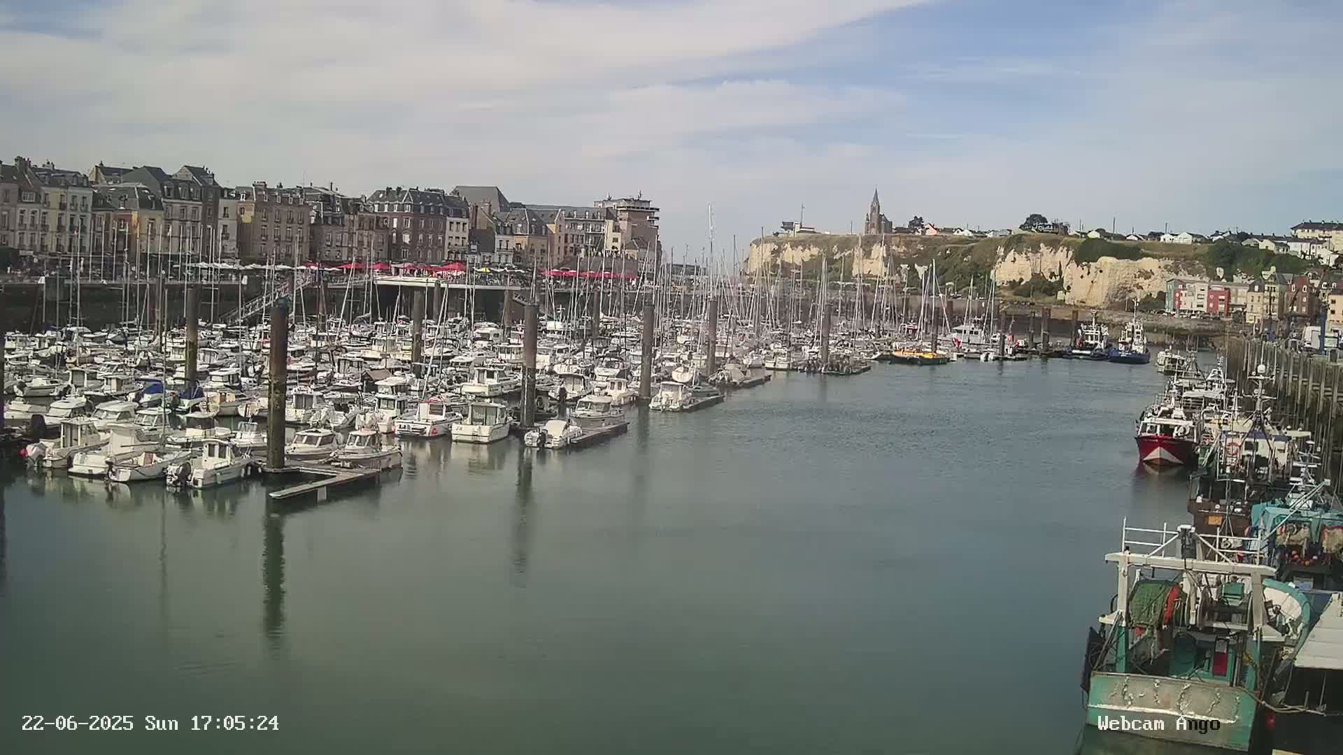 A harbor filled with numerous boats and yachts is situated in front of a town built on a cliff face under a partly cloudy sky.