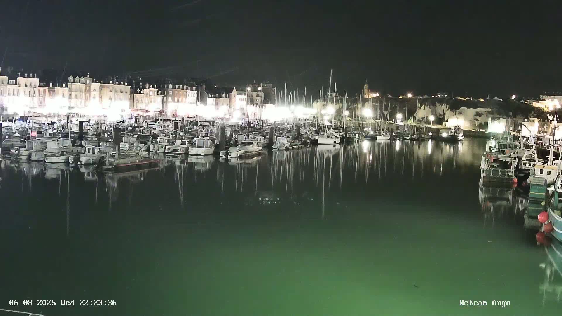 A nighttime harbor scene shows many boats moored in calm, dark-green water, reflecting the illuminated buildings and cliffs along the shoreline.