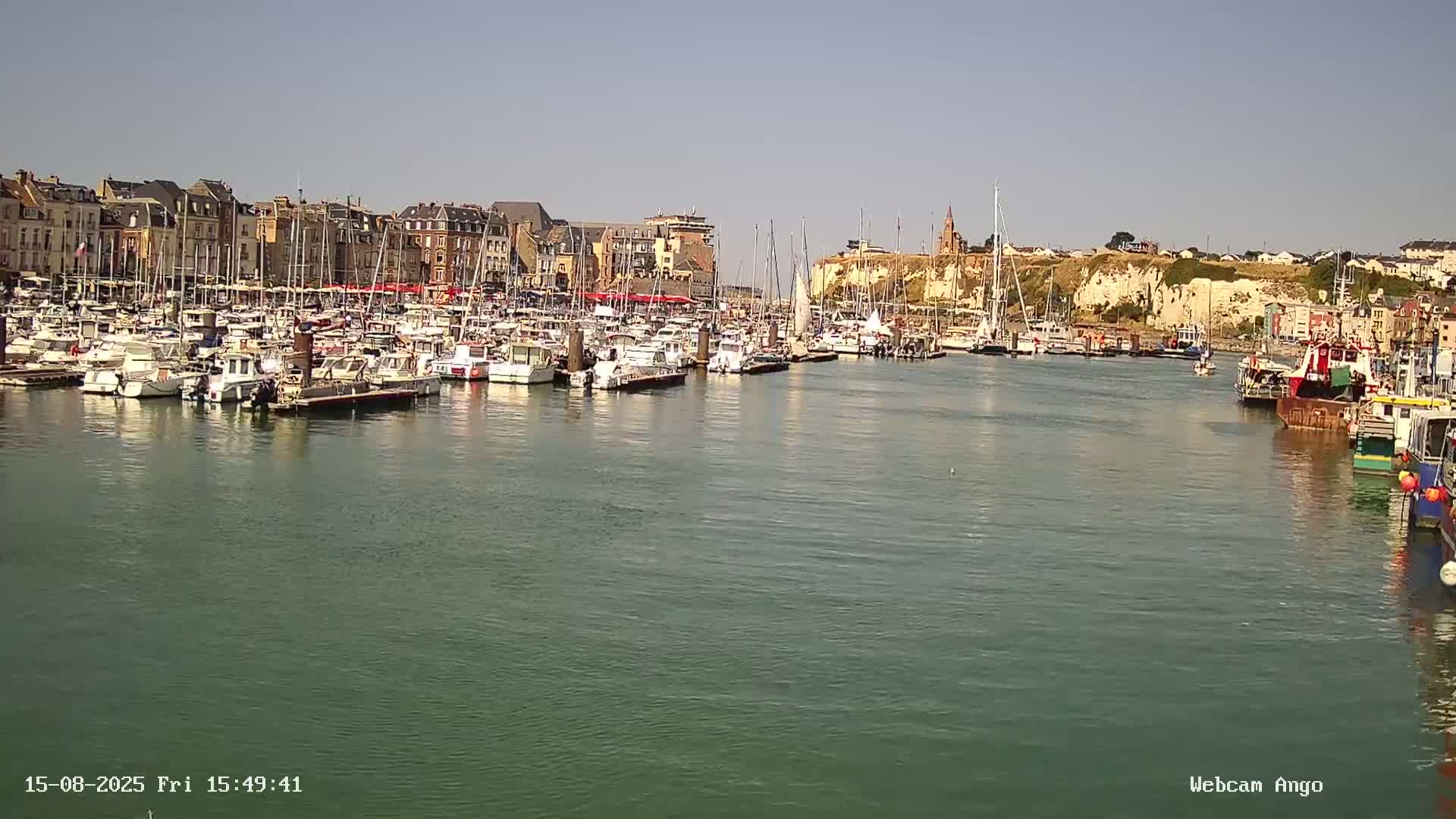 A nighttime harbor scene shows many boats moored in calm, dark-green water, reflecting the illuminated buildings and cliffs along the shoreline.
