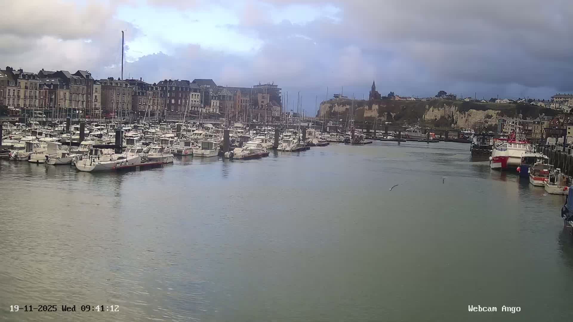 A marina packed with numerous boats of various sizes is bordered by traditional buildings on one side and a town atop a dramatic cliff with a church spire on the other, all under an overcast and cloudy sky.