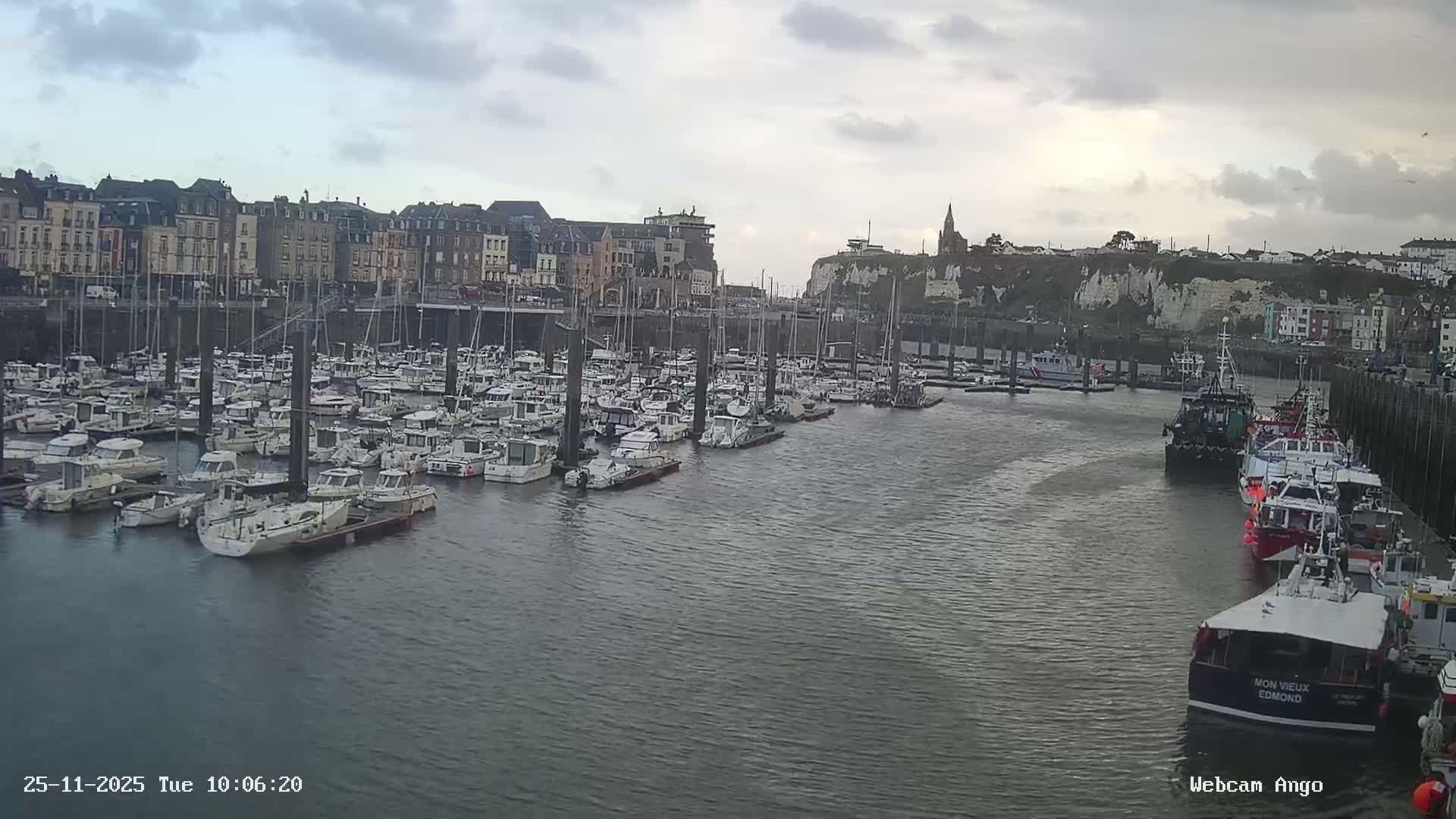 A wide view depicts a harbor crowded with numerous docked boats, flanked by a dense row of multi-story buildings on the left and white cliffs topped with a church and houses on the right, all under an overcast sky with soft, diffused light.